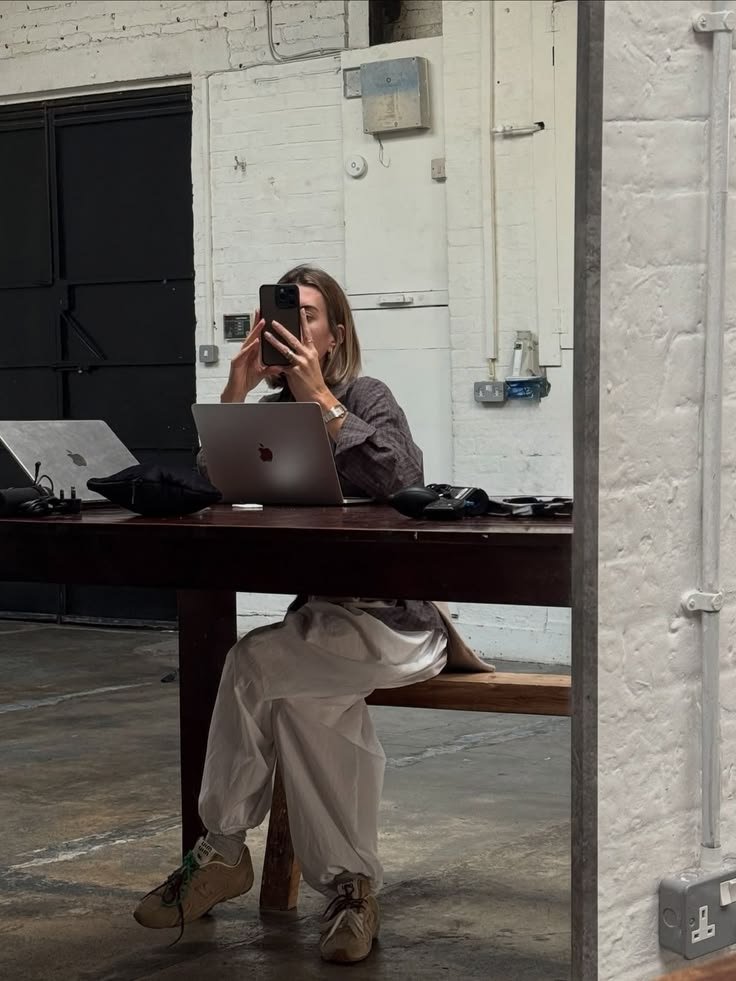 Eine Frau sitzt an einem Holztisch in einem industriellen Raum, benutzt ein Laptop und macht ein Selfie mit einem Smartphone, das sie in der linken Hand hält.