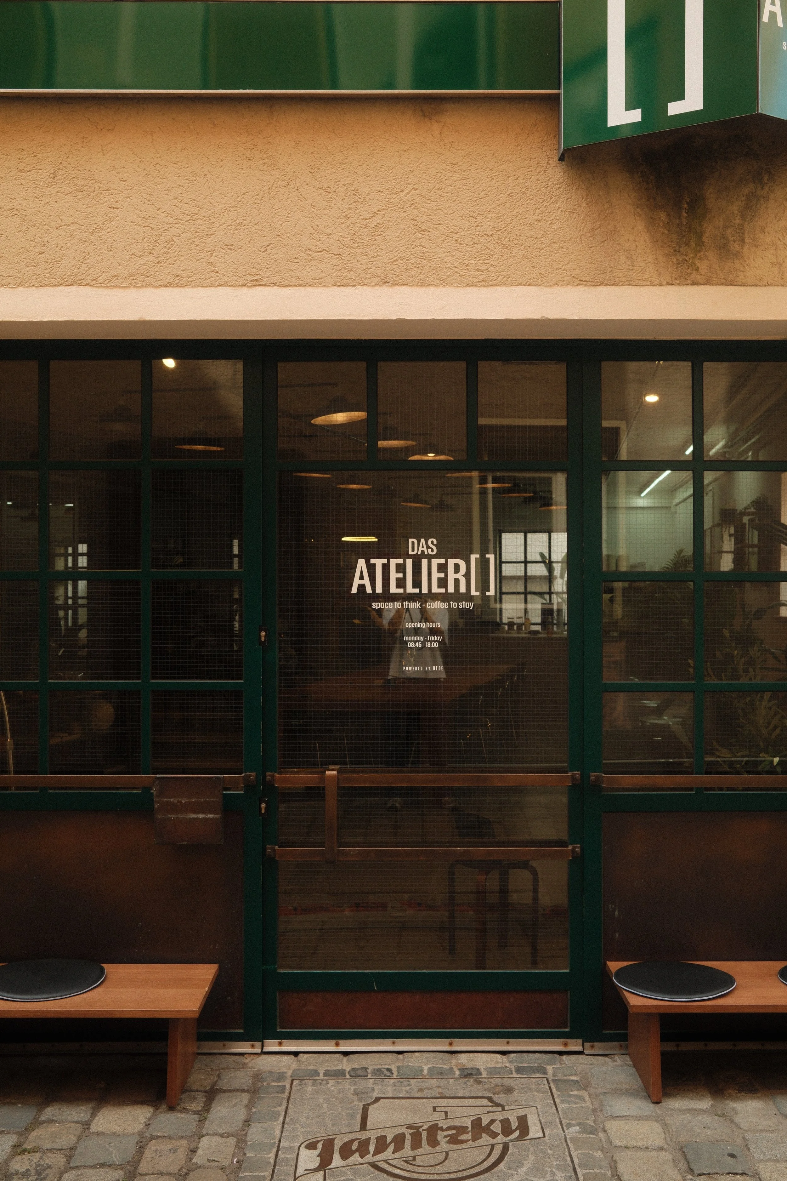 Die Tür eines Cafés namens "Das Atelier", mit einer Glasfüllung und einem Schild mit den Öffnungszeiten. Zwei kleine Holzbänke mit schwarzen Sitzkissen stehen vor der Tür, auf dem Boden liegt ein Reinigungsmatte mit dem Schriftzug "Janiczek"