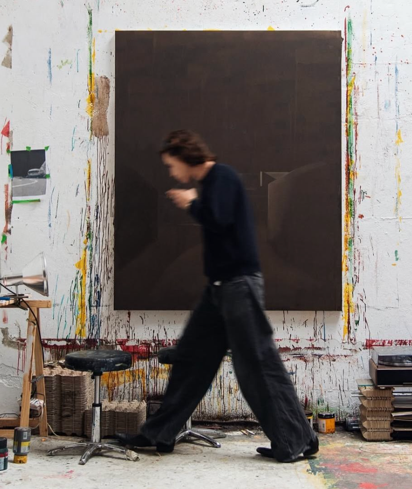Ein Mann in einem schwarzen Pullover und schwarzen Hosen läuft vor einem großen schwarzen Kunstwerk in einem Kunststudio, umgeben von bunten Farbspritzern und Malerutensilien.