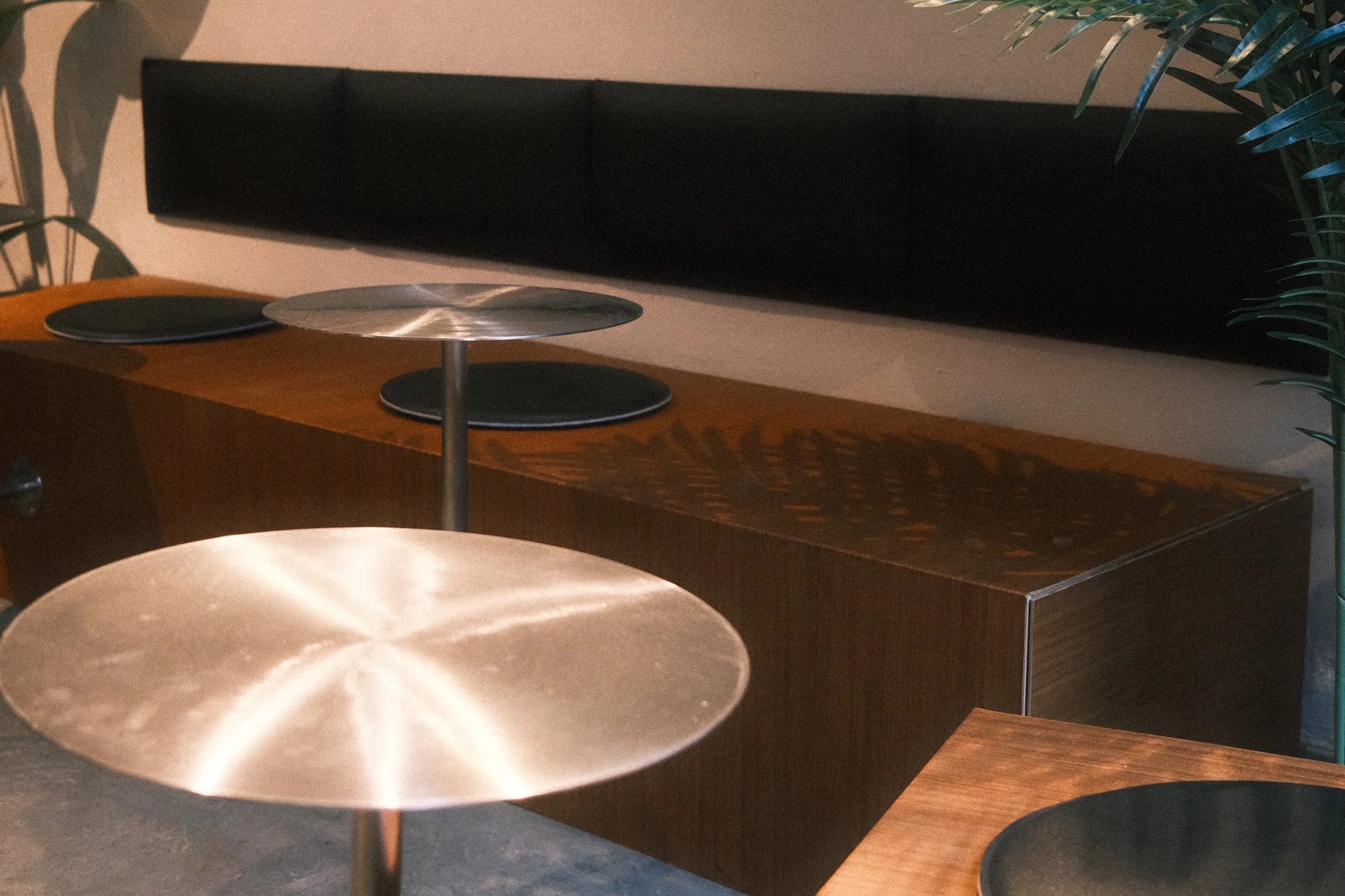 Innenaufnahme mit dunklem Sideboard, einem runden Metalltablett, drei schwarze Teller und einem Stuhl mit cremefarbenem Sitzpolster, grüne Pflanzen im Hintergrund.