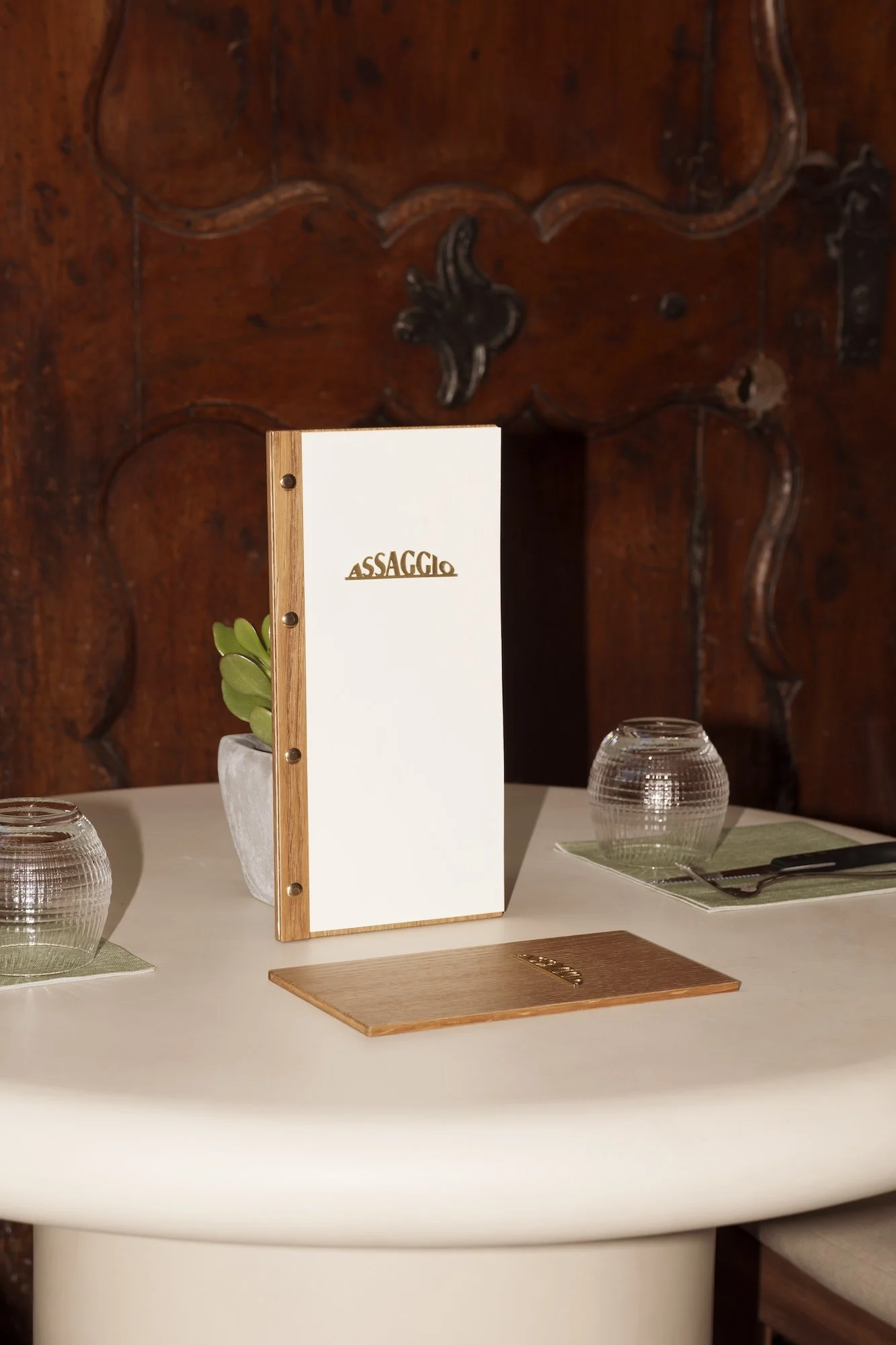 Assaggio restaurant menu on a dining table with glass candleholders, utensils, and a potted plant against an ornate wood backdrop