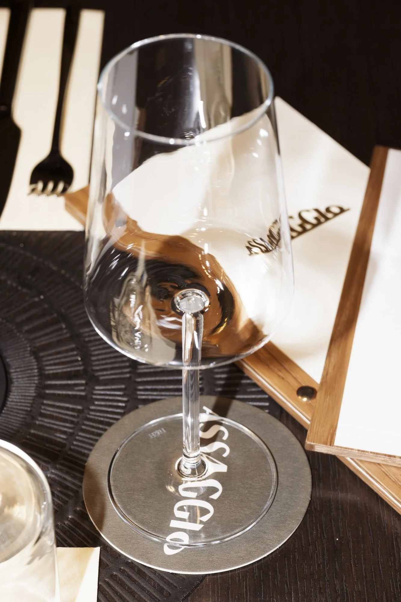 Assaggio Merano - Empty wine glass on a branded coaster at a restaurant table with white napkins and black cutlery