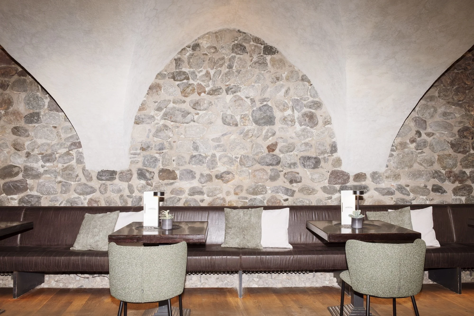Rustic restaurant dining area with stone walls from the 17th century, leather banquette seating, wooden tables, upholstered chairs, lamps, and pillows