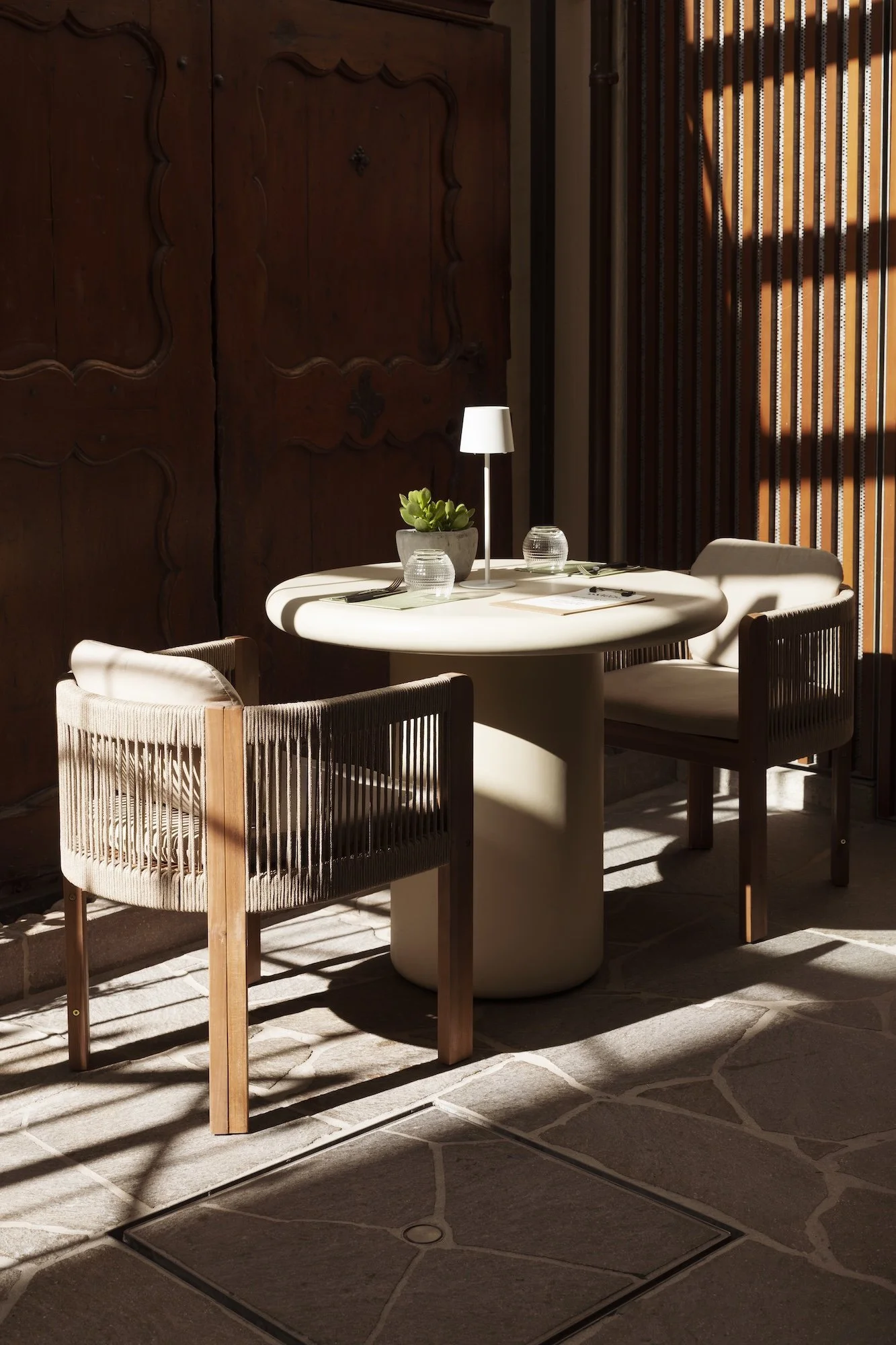 Elegant dining area with a round table, two woven armchairs, a lamp, succulent, and sunlight through vertical blinds.