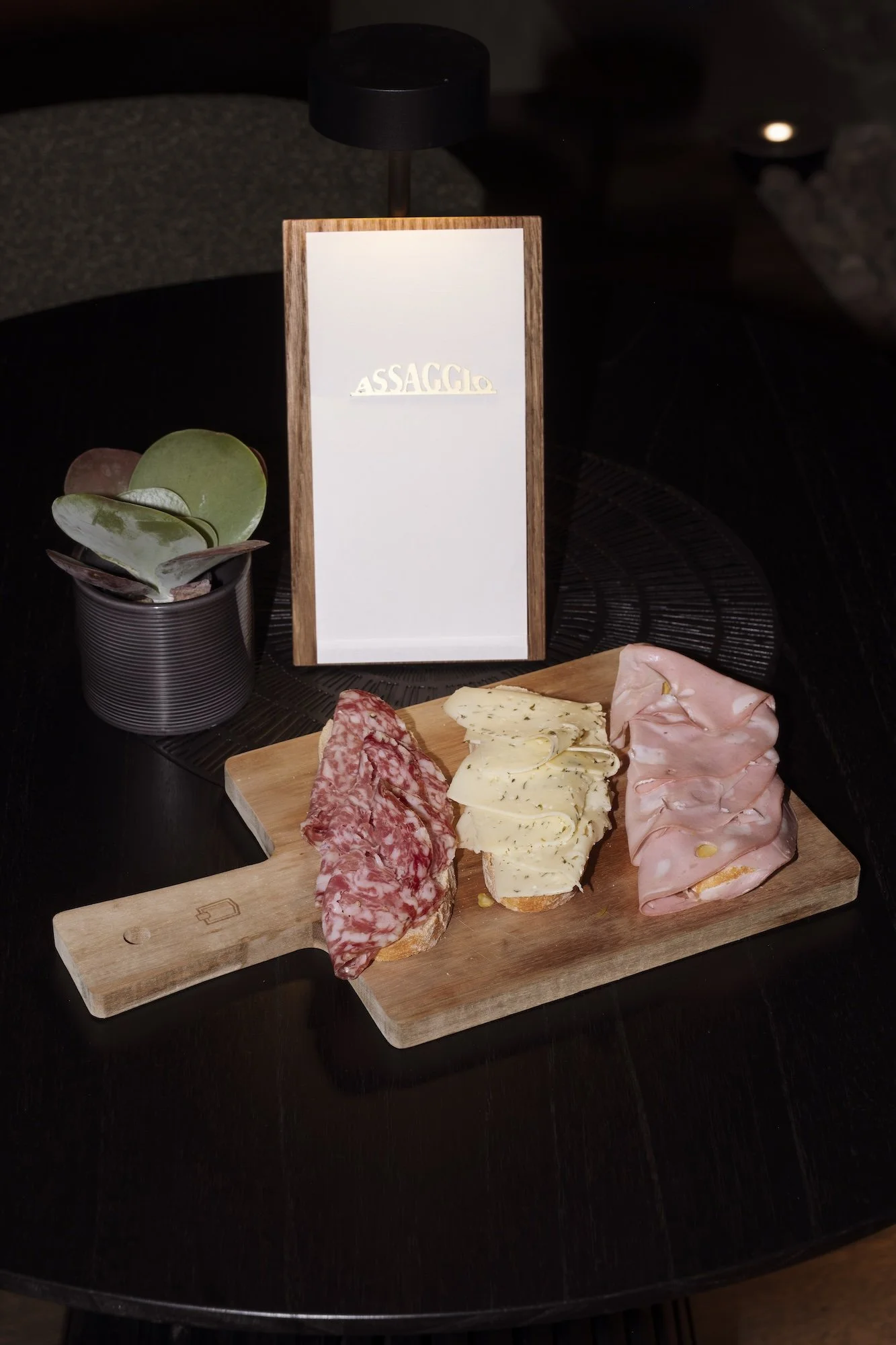 Charcuterie-style sandwich bites with salami, herb cheese, and ham on a wooden serving board beside a potted succulent and menu labeled Assaggio.