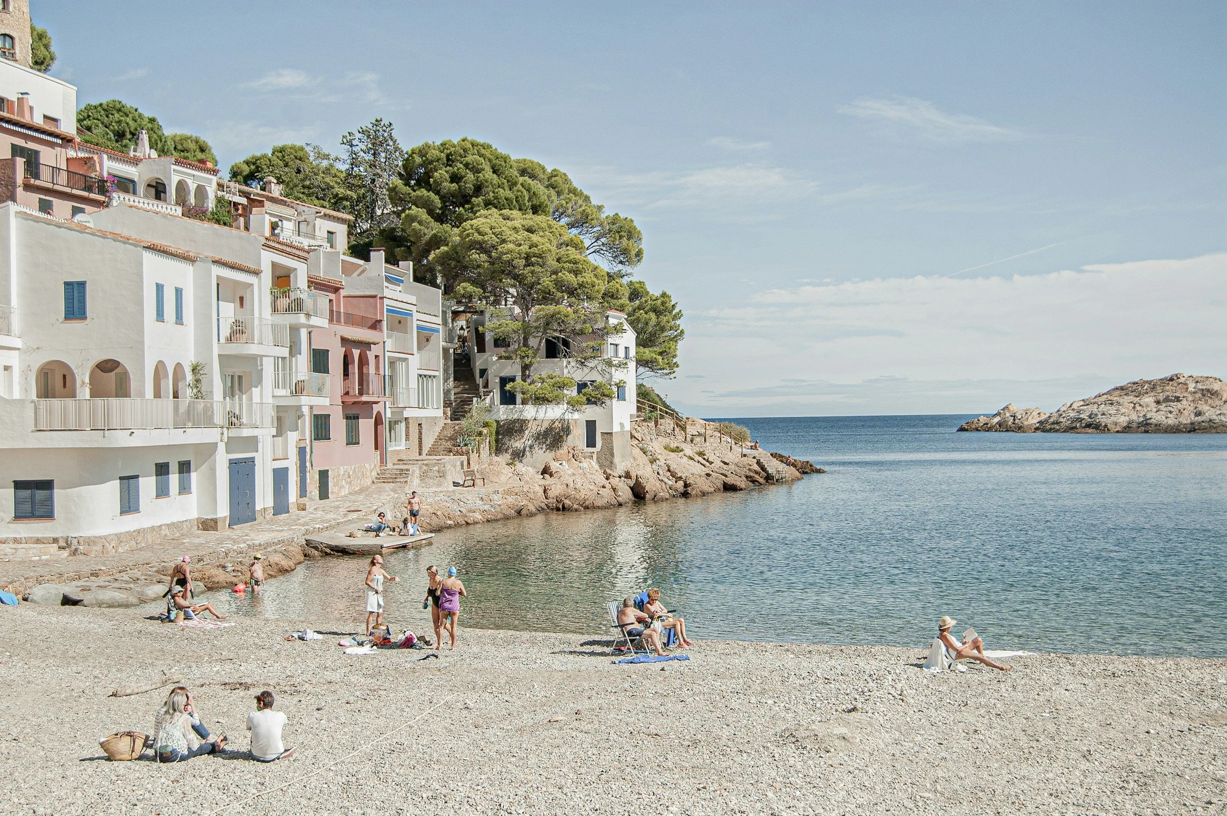 People relaxing on a pebble beach near the sea, with colorful seaside buildings and green trees on a hillside in the background under a blue sky.