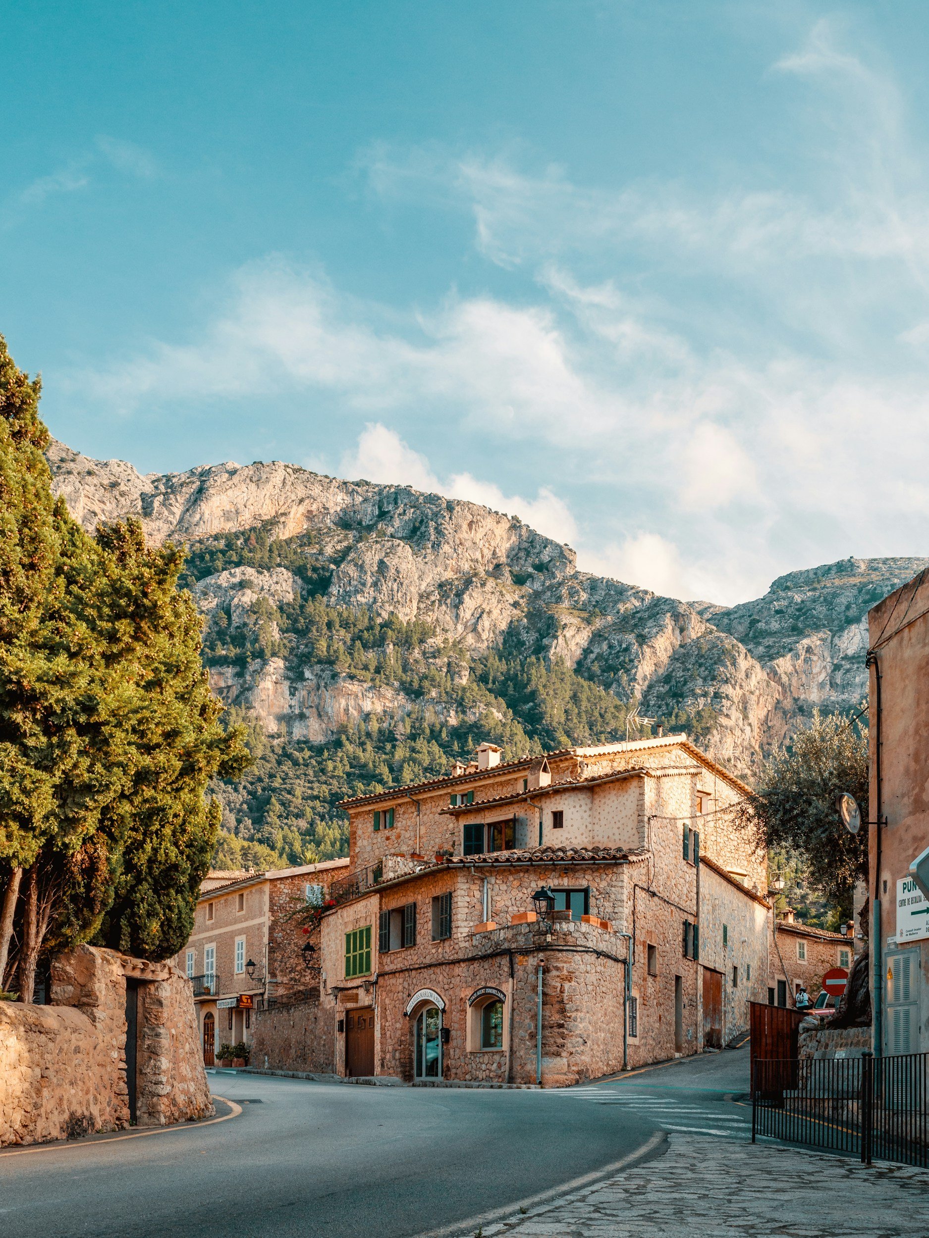 A small European village with stone buildings lining a curved street, nestled against a mountainous backdrop under a partly cloudy sky.