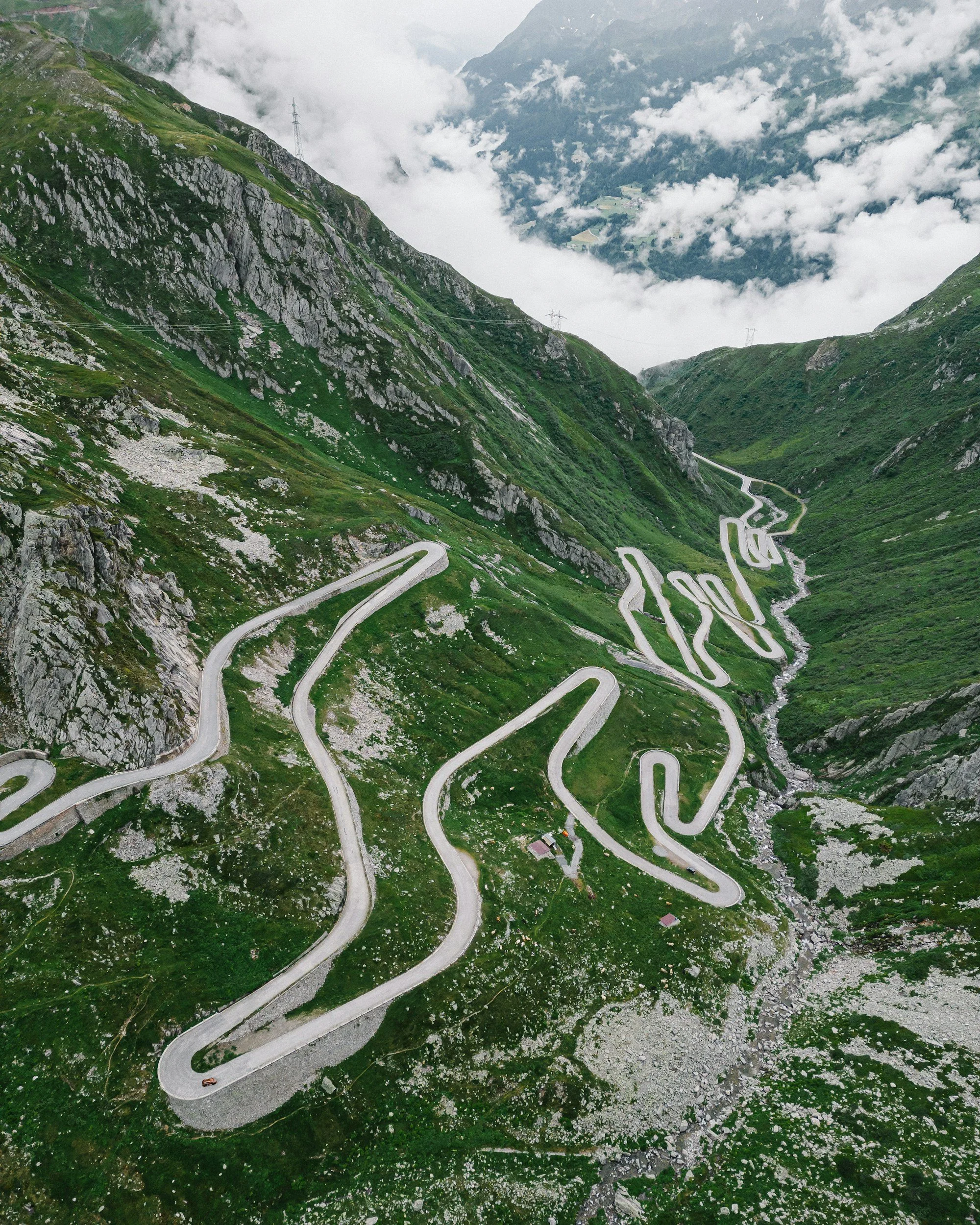 Aerial view of a winding mountain road cutting through lush green hillsides with rocky terrain and a small vehicle traveling along it.