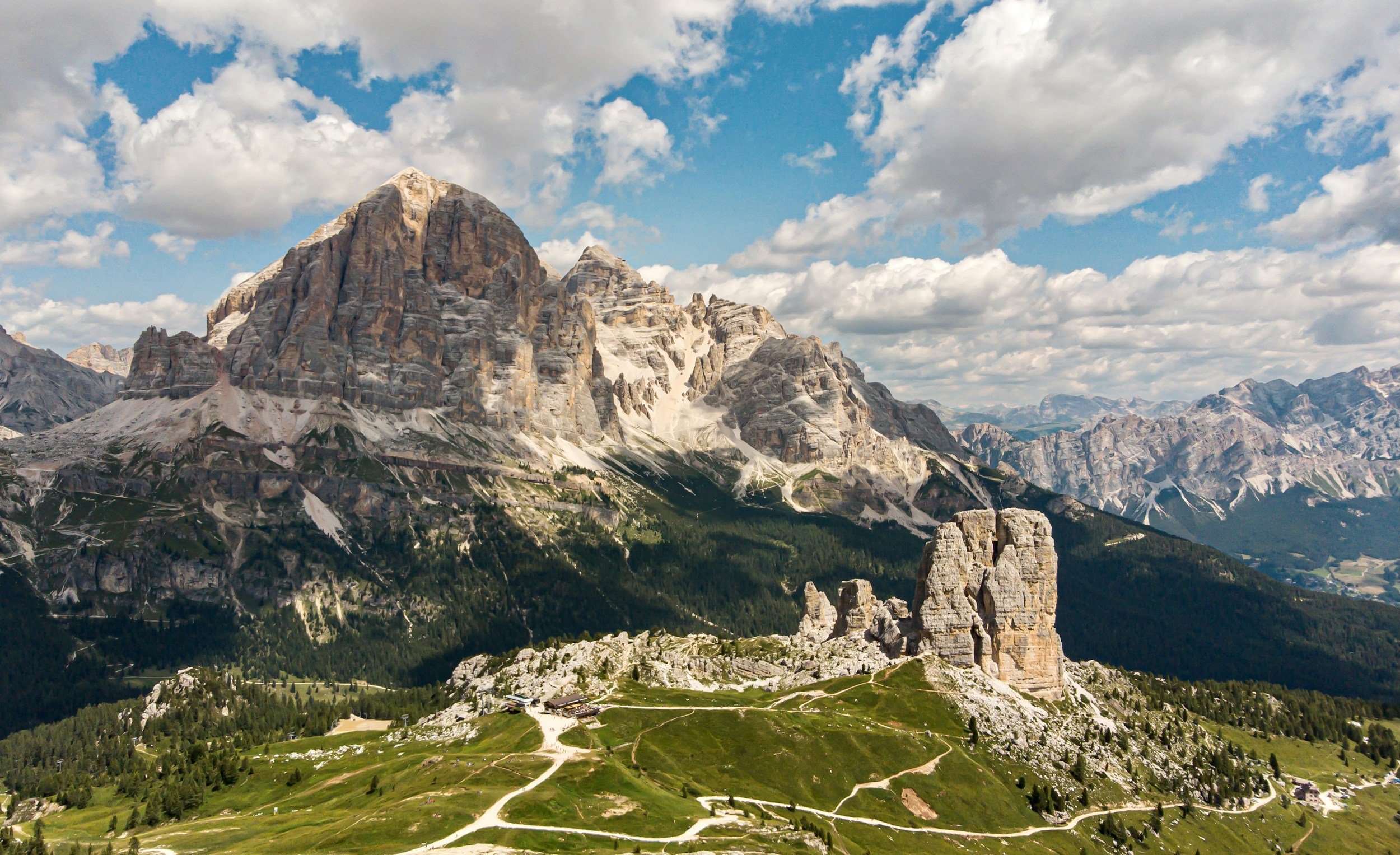 A mountain range with towering rocky peaks, some snow-capped, under a partly cloudy sky, with green valleys and pathways in the foreground.