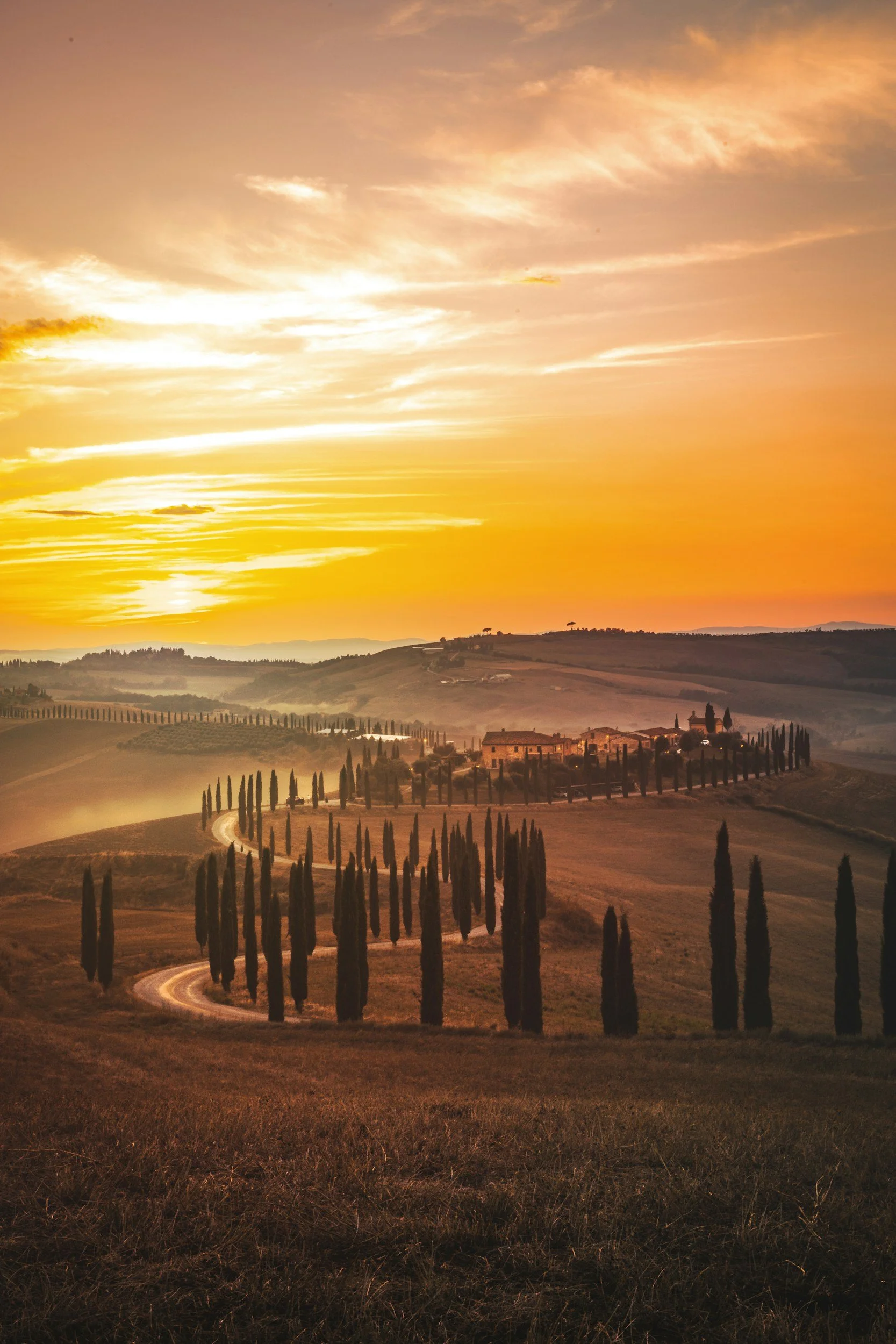 A scenic sunset over the countryside in Tuscany, Italy, showing a winding road lined with tall cypress trees, rolling hills, and a distant house with trees, under a colorful sky with orange, pink, and yellow hues.