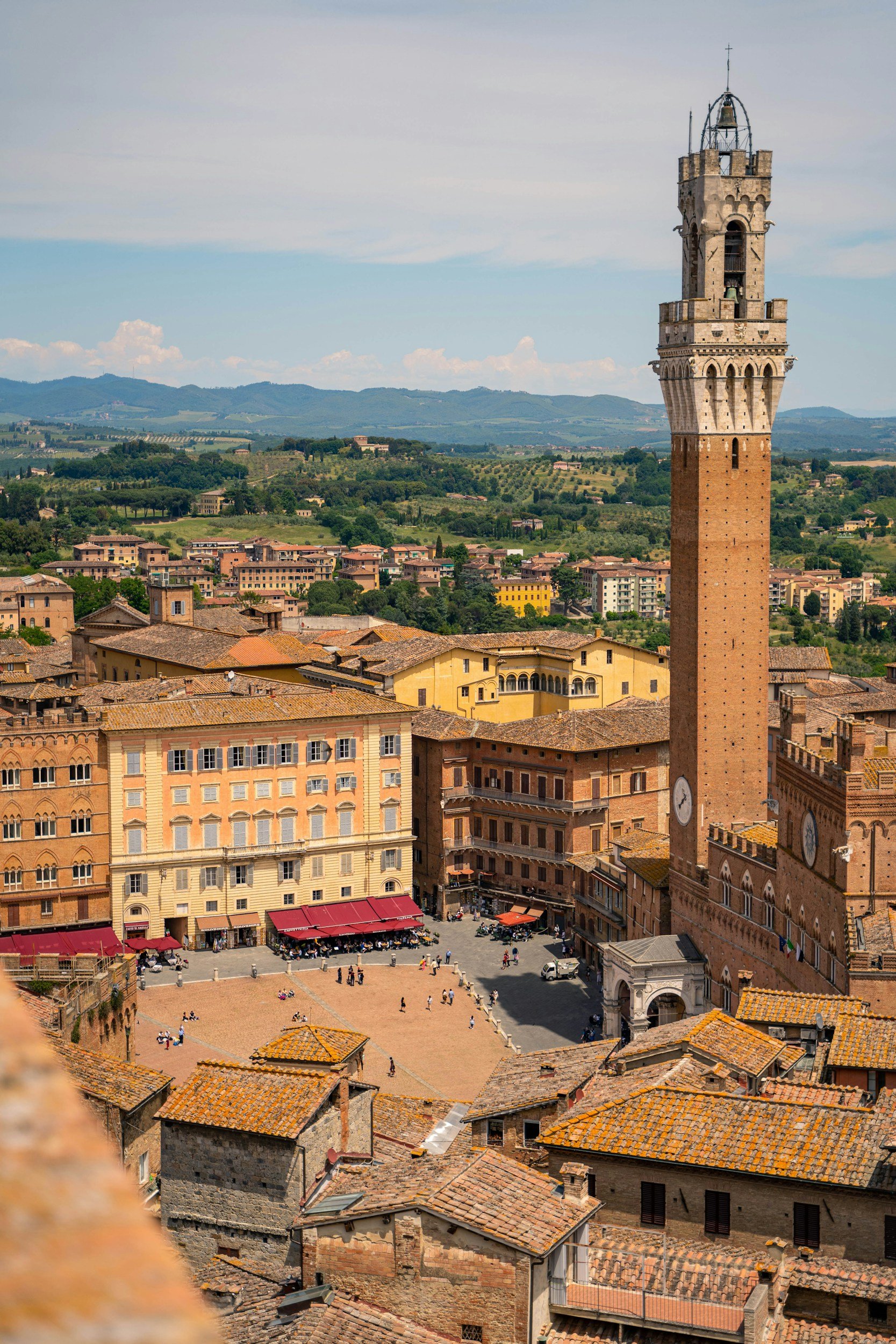 Overview of an Italian historic town with a tall clock tower, terracotta roofs, and a central plaza with people, against rolling green hills under a blue sky with some clouds.