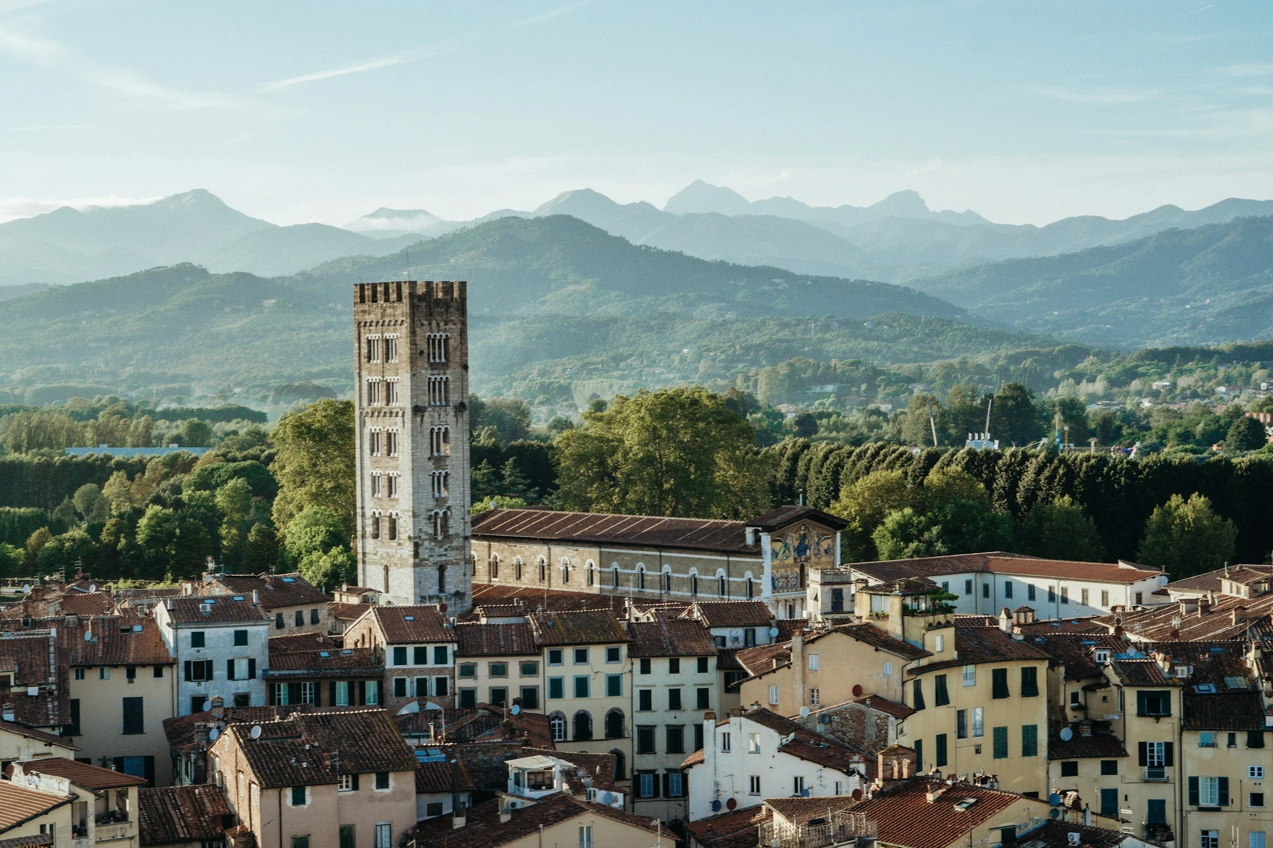 A scenic view of a historic town with a tall stone church tower, surrounded by houses with red-tiled roofs and green trees, set against rolling hills and mountains in the background.