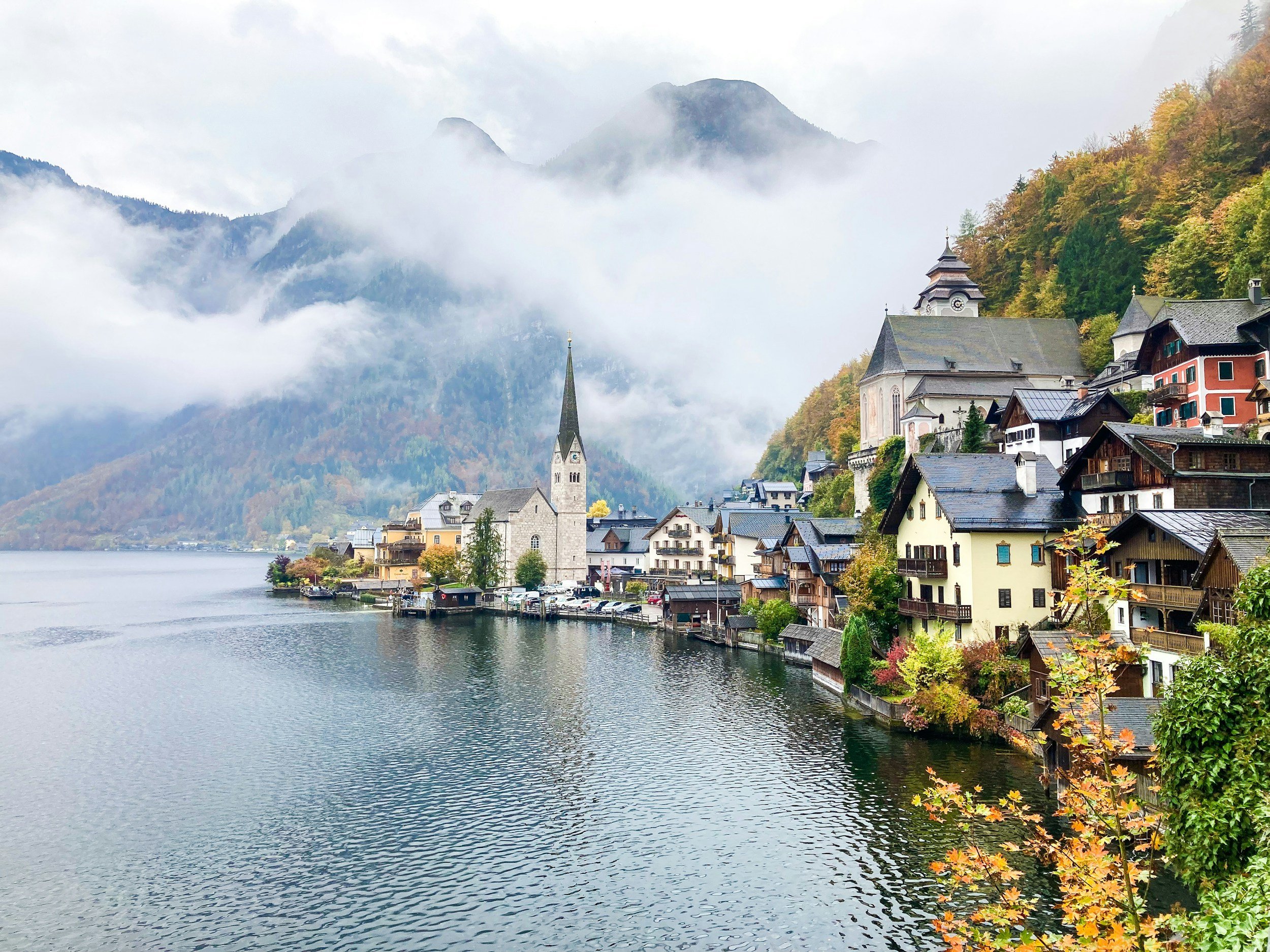 A lakeside village in the mountains with colorful autumn foliage, a church steeple, and misty mountains in the background.