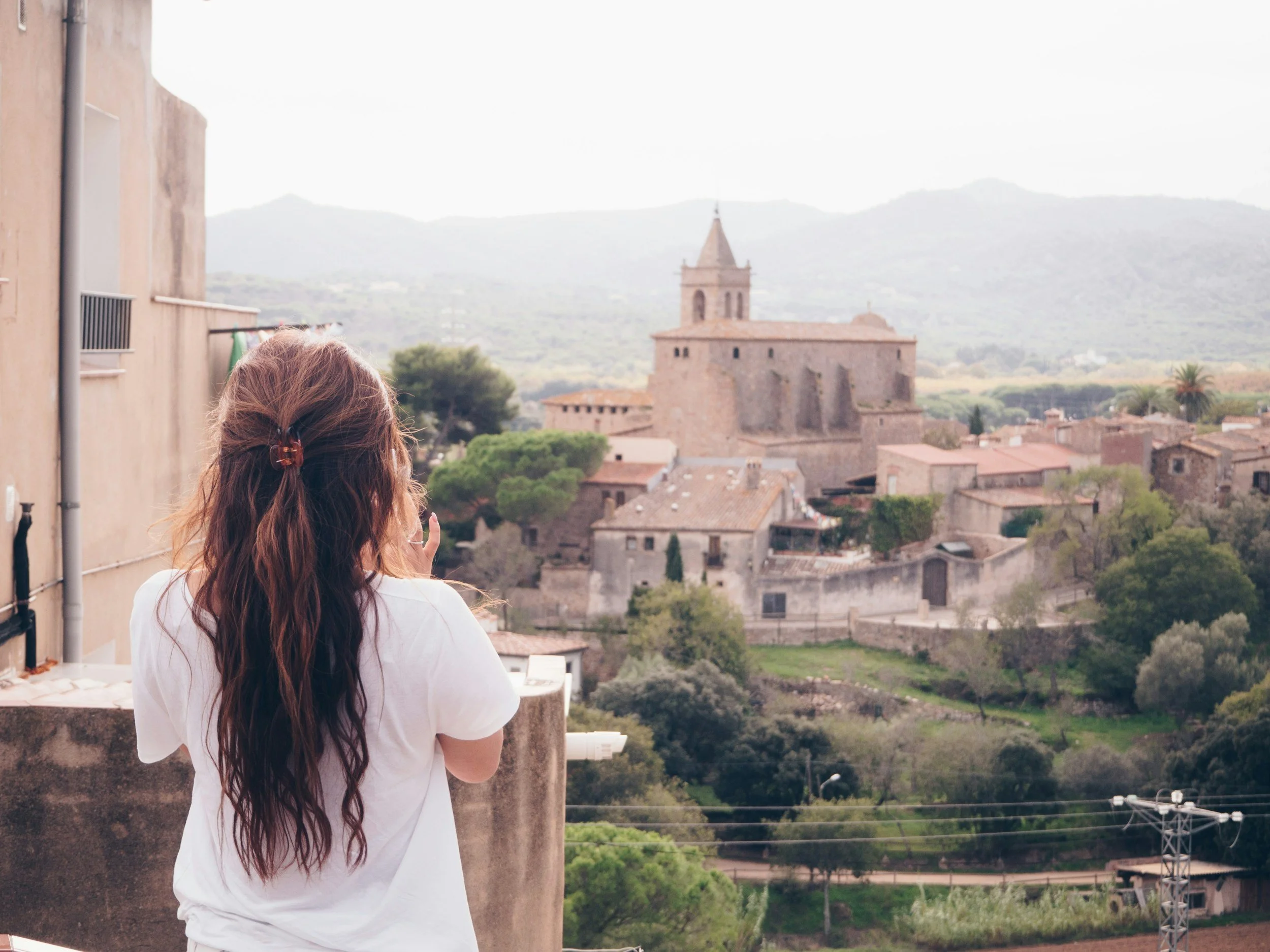 A woman with long, wavy hair, seen from behind, taking a photo of a historic town with a church tower in the background. The town is surrounded by green hills and mountains.