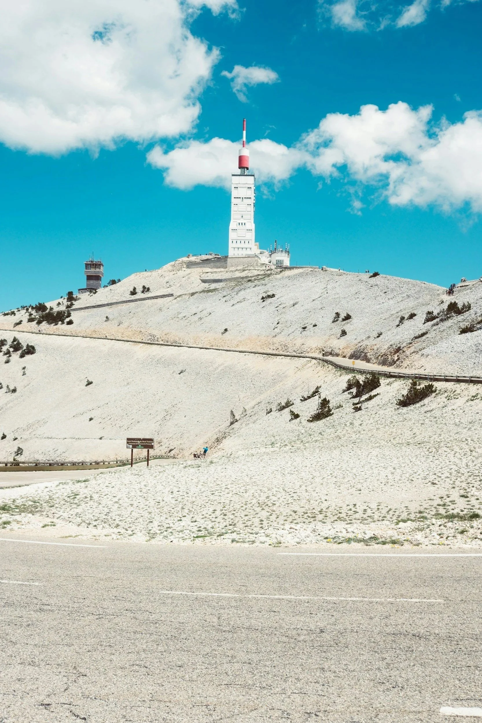 A tall white building with a red and white antenna on top, situated on a mountain with sparse vegetation, under a blue sky with scattered clouds.