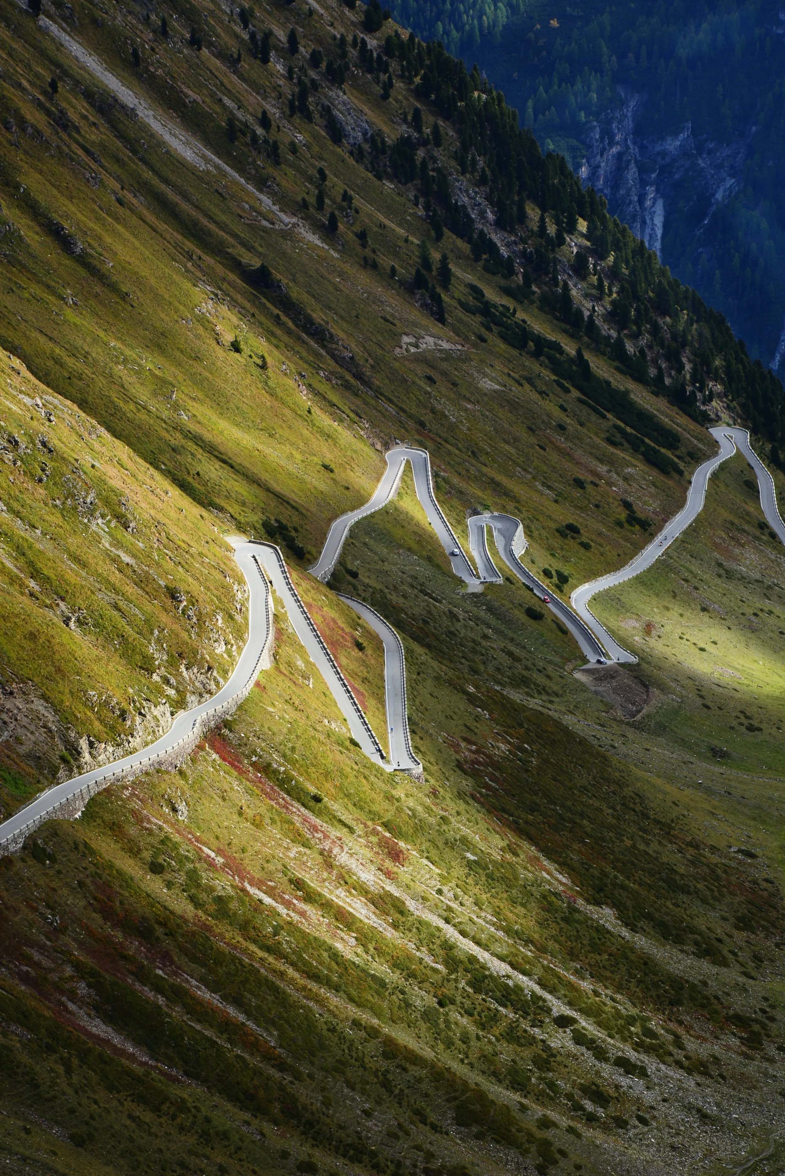 A winding mountain road with sharp turns zigzags down a steep hillside covered in green vegetation and some rocks, with forested mountains in the background.