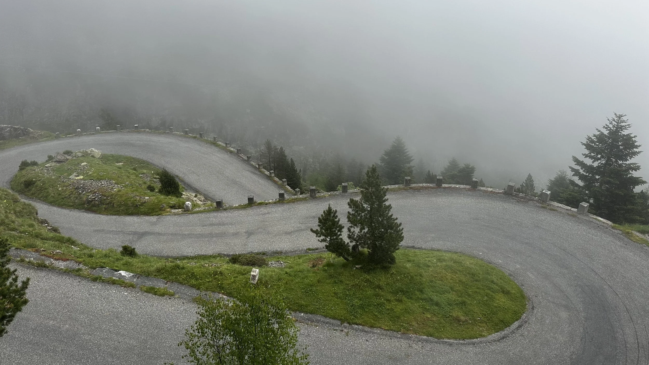 A winding mountain road with sharp curves, lined with small white barriers, amidst green grass and trees, shrouded in fog.