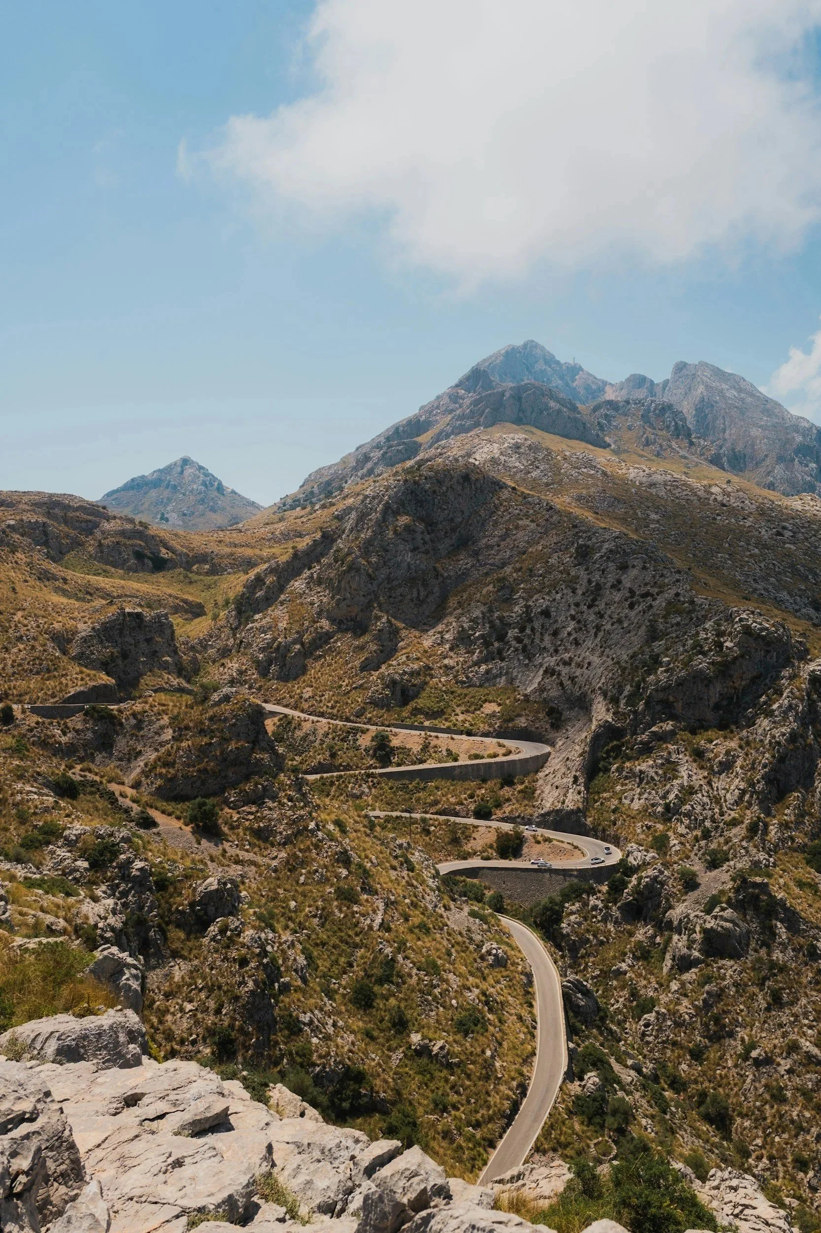 A winding mountain road with cars, surrounded by rocky hills and sparse vegetation, under a partly cloudy sky.