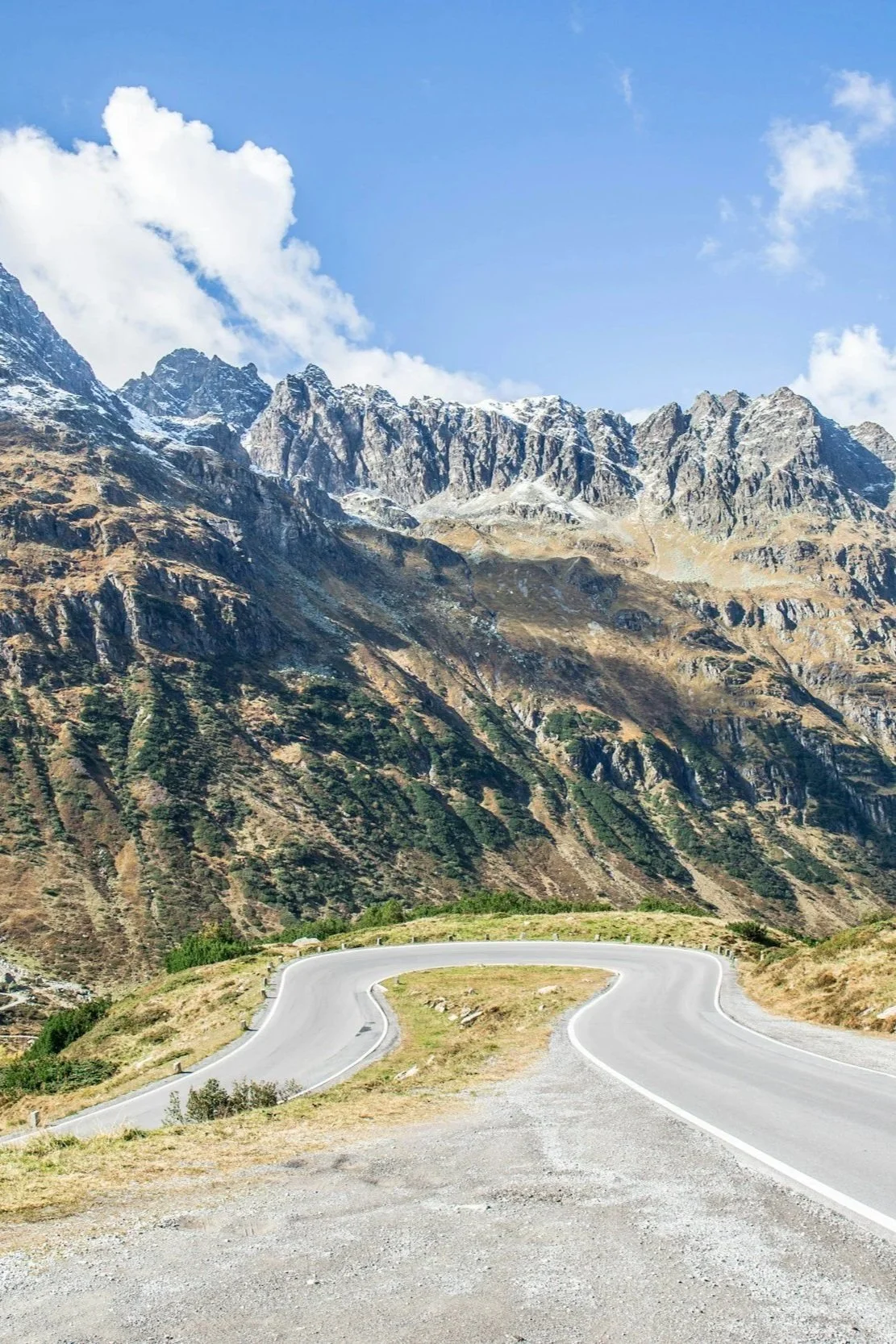 A winding mountain road at the base of tall, rugged, snow-capped peaks under a partly cloudy blue sky.