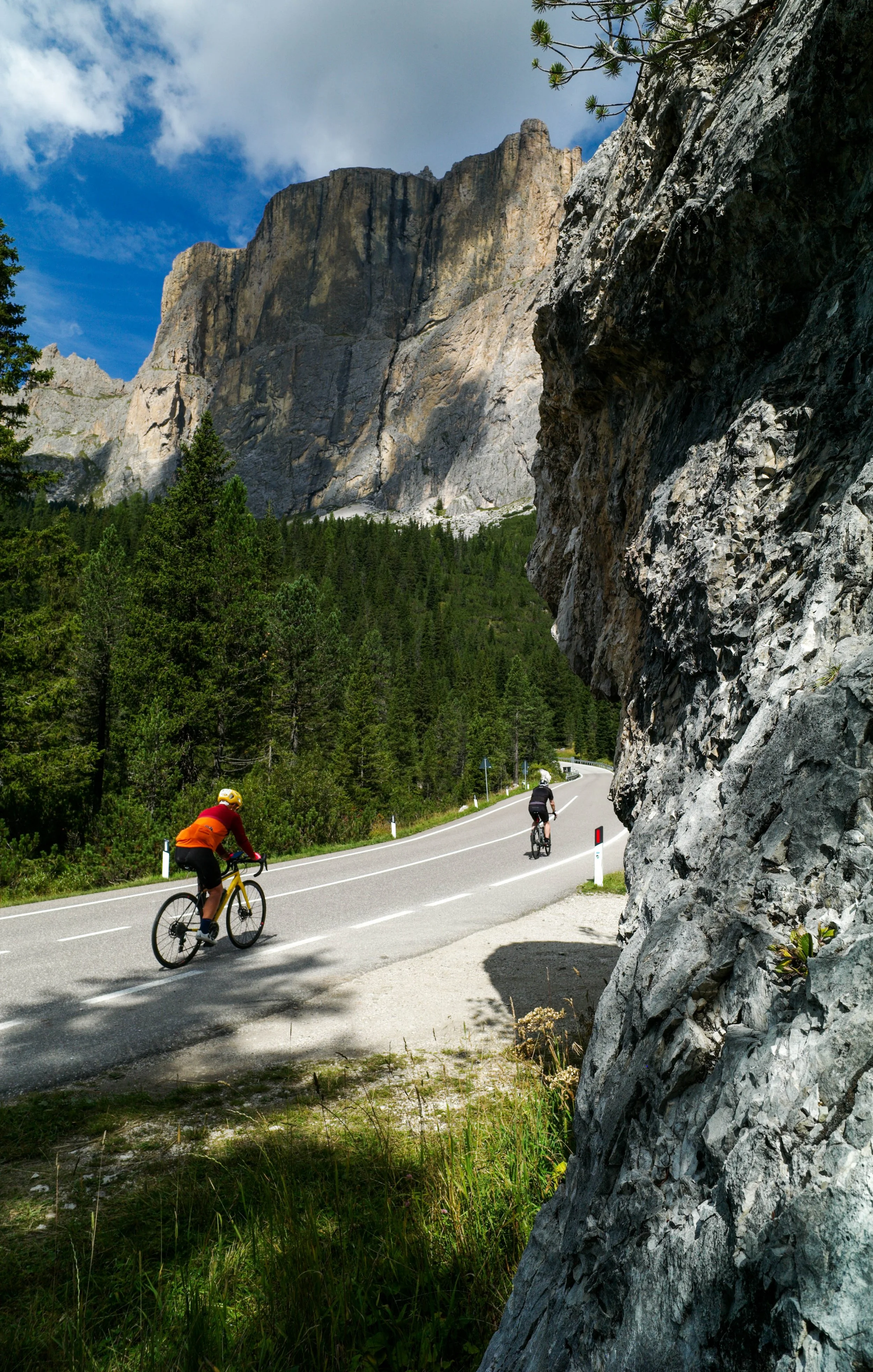 Cyclists on a winding mountain road surrounded by tall rocky cliffs and dense green trees, with a partly cloudy sky overhead.