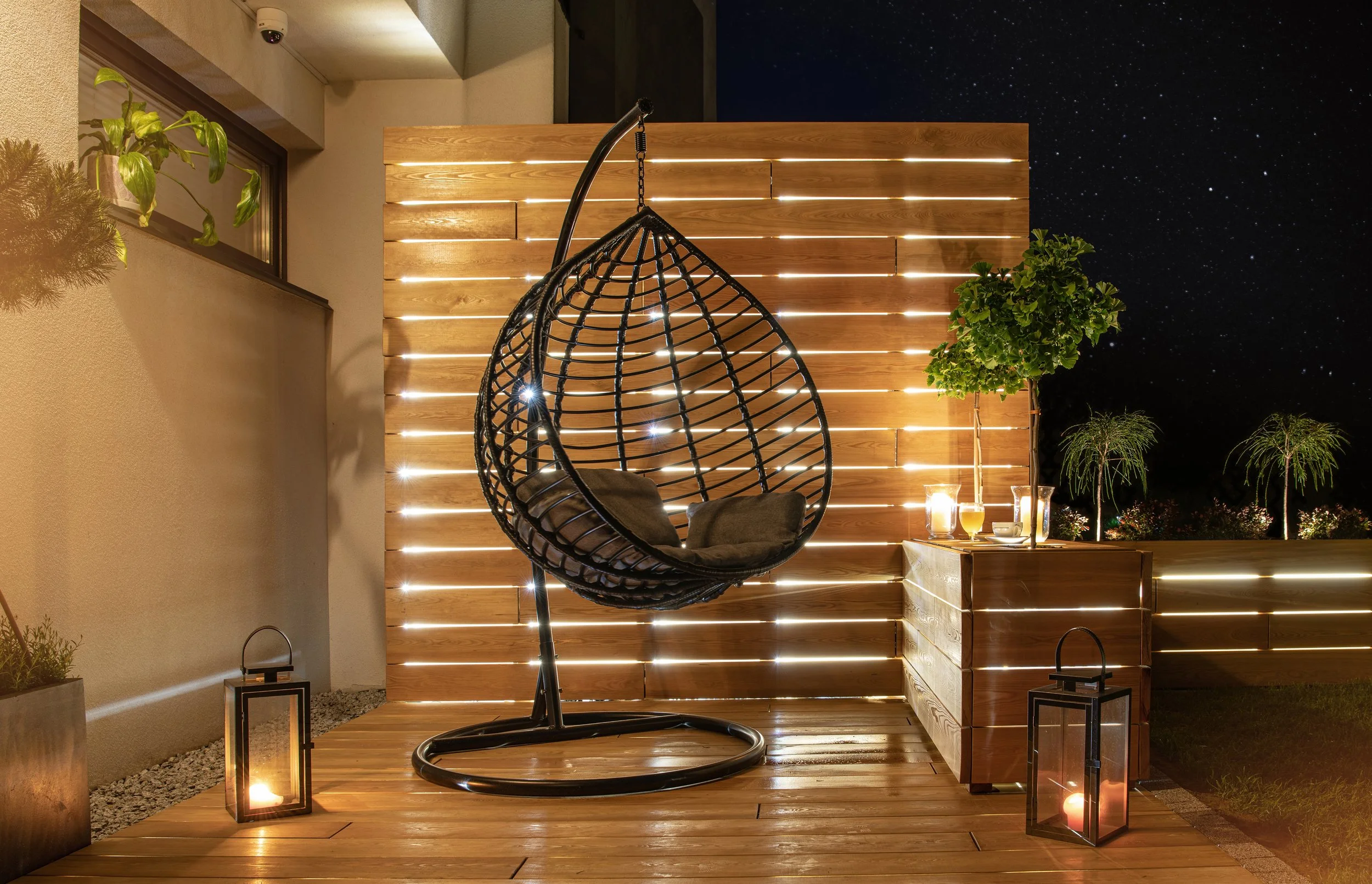 Nighttime patio scene with hanging egg-shaped swing chair, potted plants, lanterns, candles, and a wooden privacy fence illuminated by integrated lighting, under a starry sky.