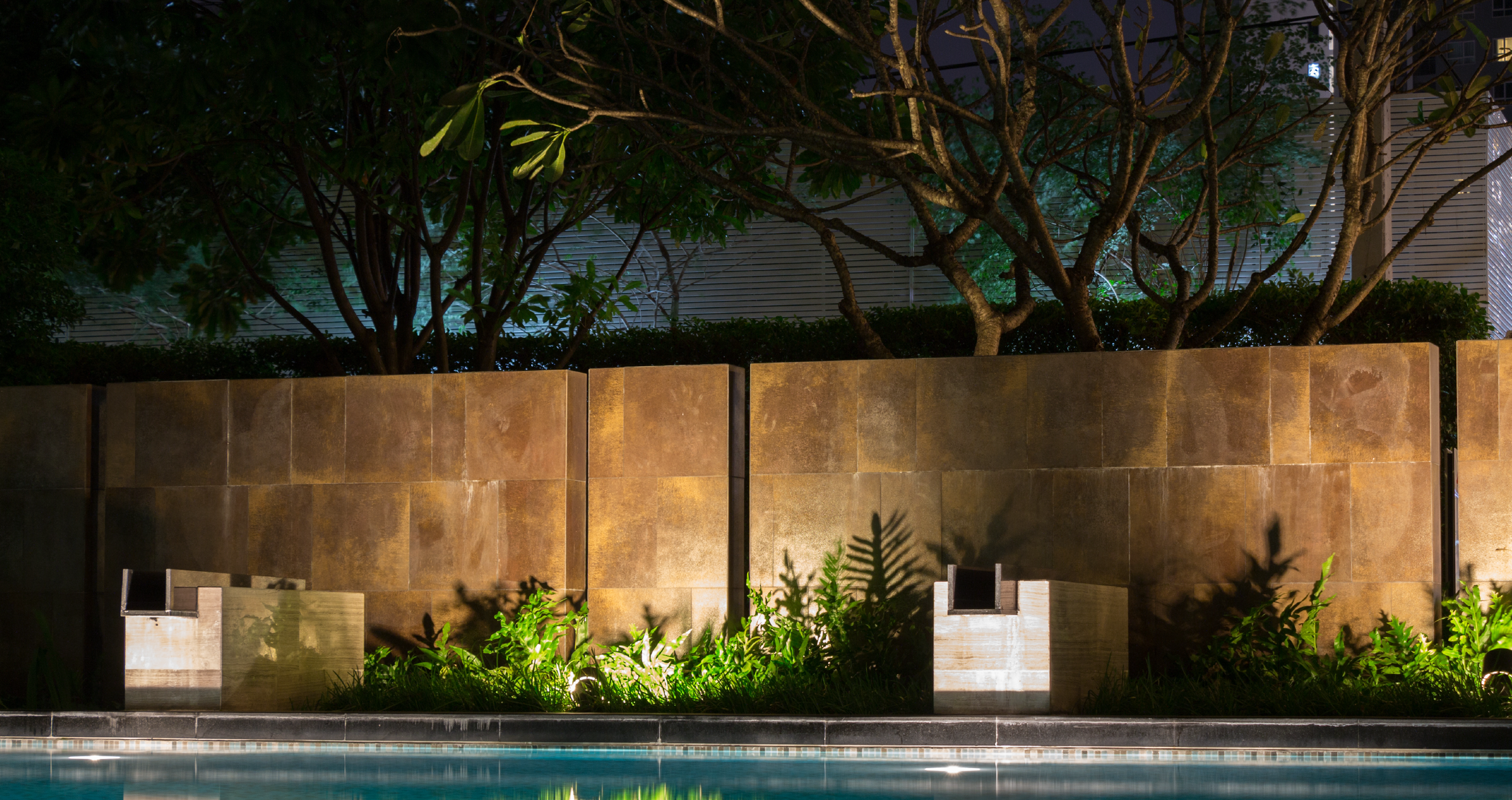 Nighttime scene of a luxury swimming pool area with a brown tiled wall, lush green plants, and trees illuminated from below.