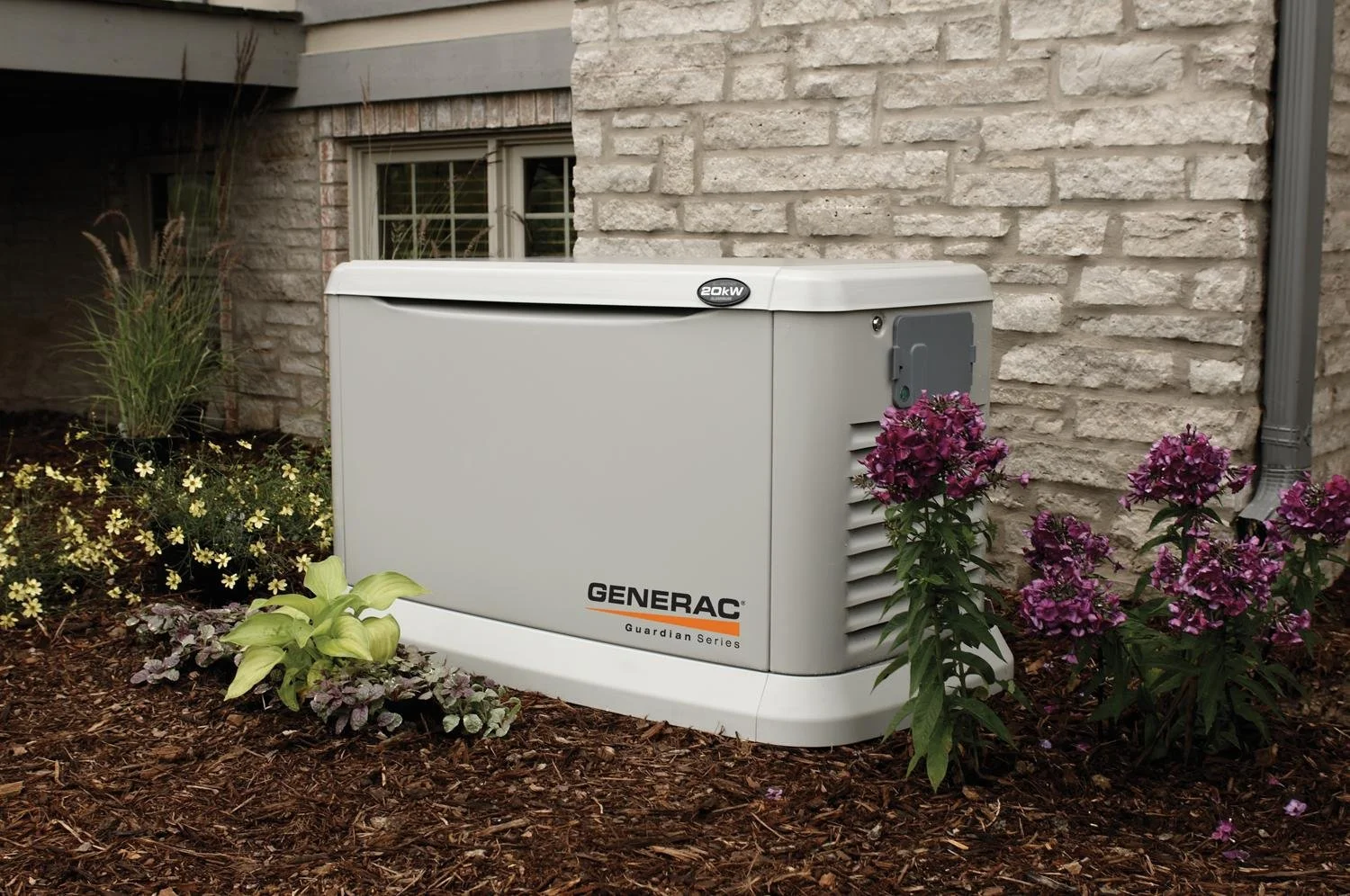 A Generac Guardian Series generator outdoors near a brick house wall, surrounded by purple and green flowering plants and mulch, under a cloudy sky.