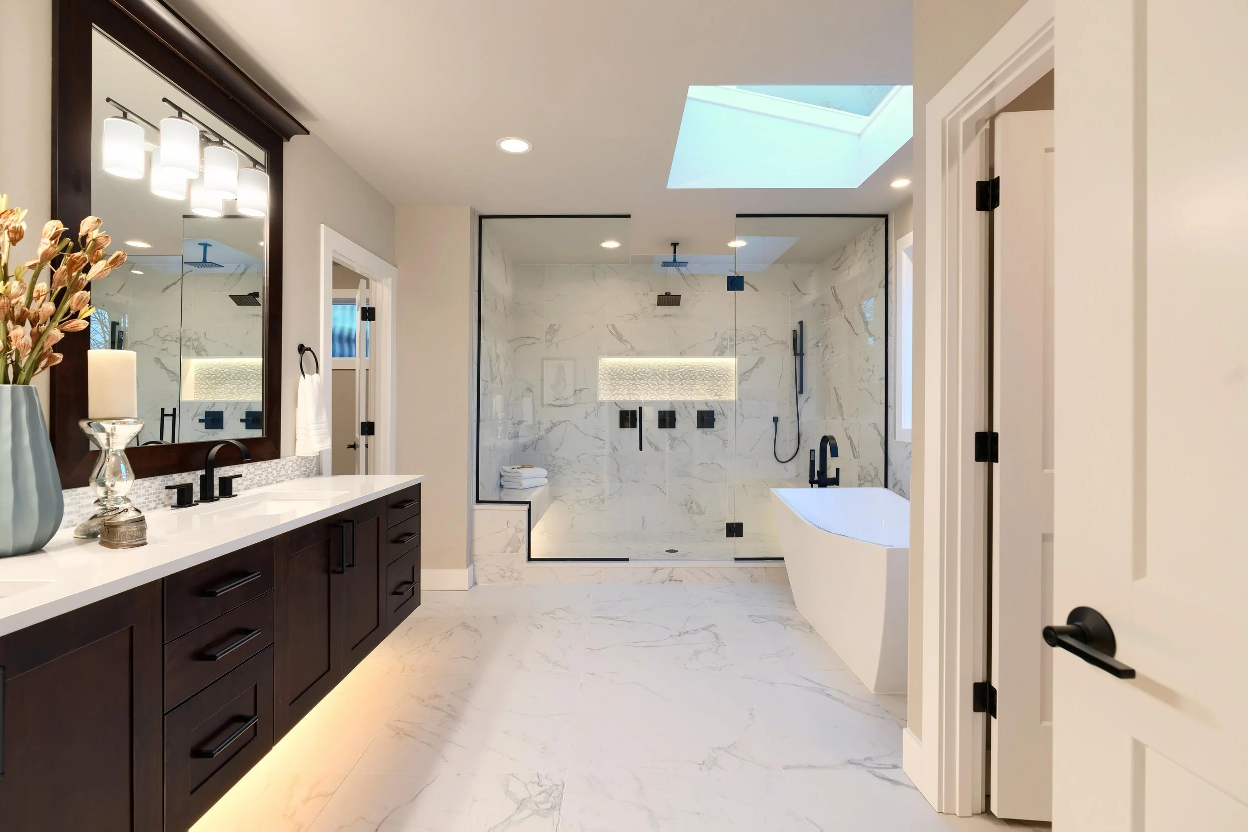 Modern bathroom with a walk-in shower with glass walls, white marble tiles, a skylight, dark fixtures, vanity with a mirror, and decorative flowers on the counter.