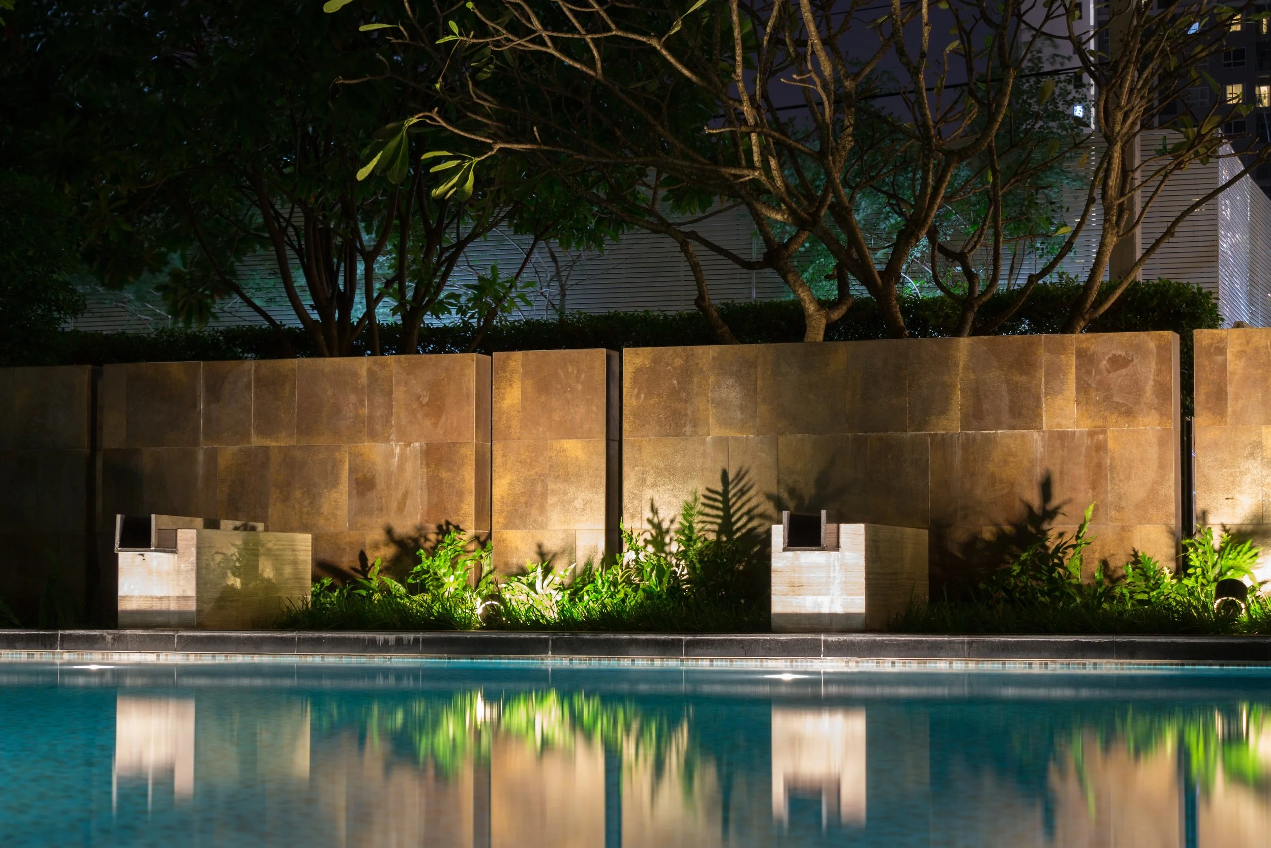 Nighttime view of a swimming pool with a brown tiled wall, illuminated landscaping, and trees in the background.