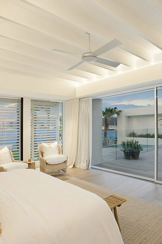 A bright, modern indoor living room with large glass sliding doors leading to an outdoor patio. The room has white walls and ceiling with a ceiling fan, and contains a bed with white linens and textured pillows, and two wicker chairs. The outdoor area features lounge chairs, potted plants, and a view of palm trees and a body of water.