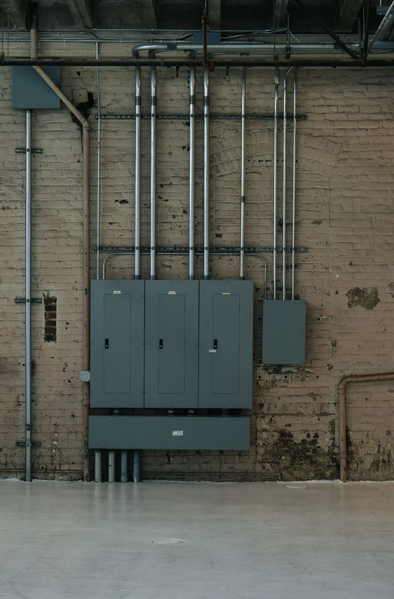 Electrical panels, pipes, and wiring mounted on a brick wall interior.