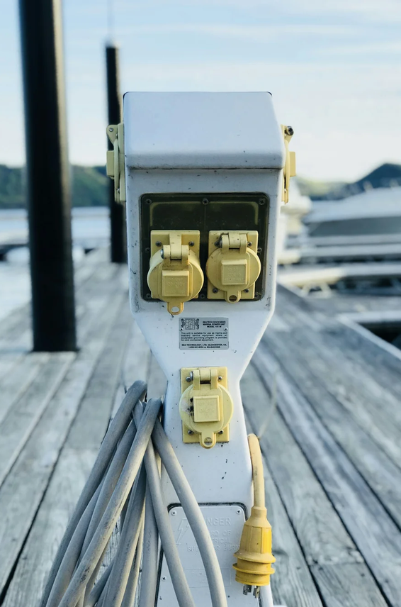 Electrical outlets and cords attached to a white post on a wooden dock with boats and water in the background