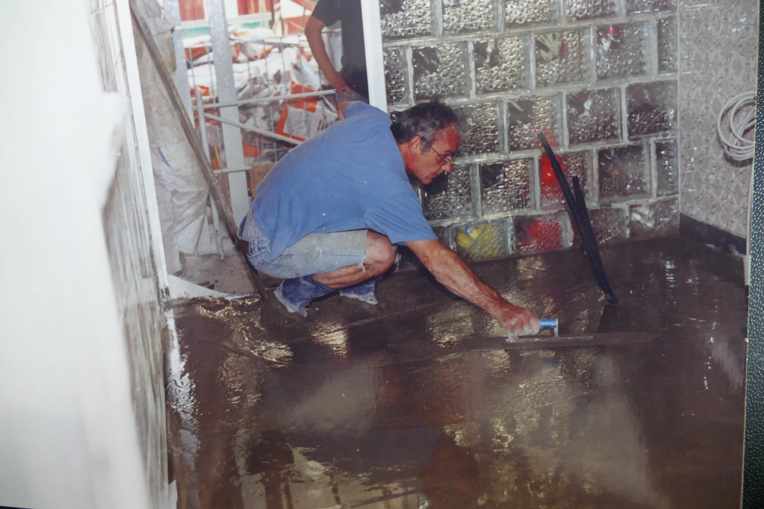 Ein Mann ist beim Fliesenlegen auf dem Boden an einer Wand. Er trägt eine blaue Arbeitshose und einen blauen T-Shirt. Der Raum wirkt noch im Bau und hat eine Wand mit Glassteinen. Es liegen Werkzeuge und Baumaterialien im Raum.