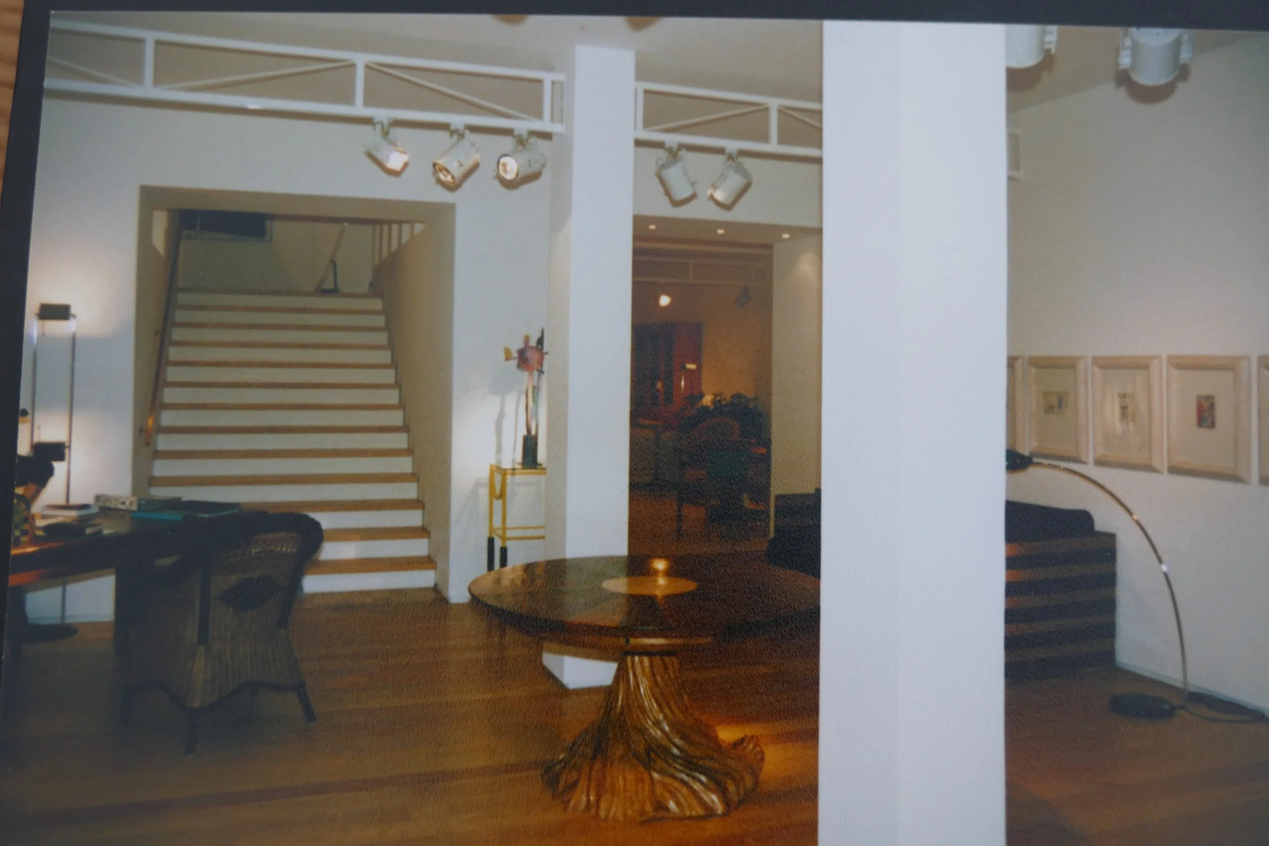 Innenaufnahme eines Wohnzimmers mit Treppe, Runden Esstisch, Stühlen und Stehlampe, gebäudeartige Raumgestaltung mit weißen Wänden und Holzfußboden.