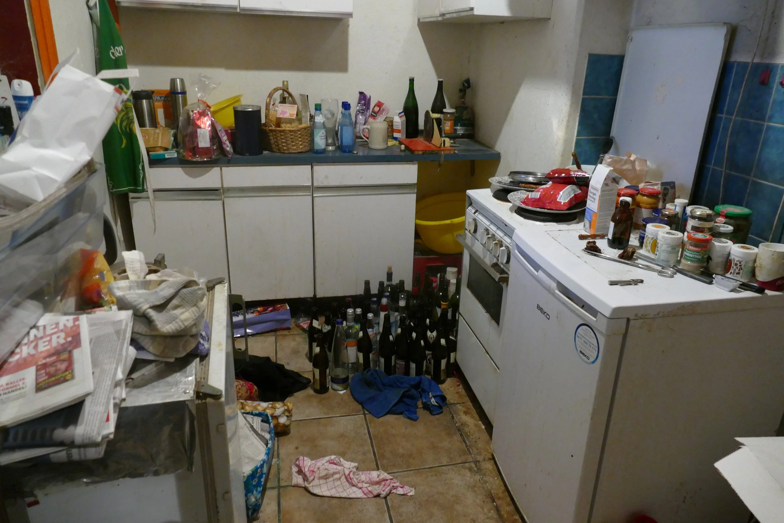 Un unordentlicher Küchenkeller mit abgelegenen Flächen, vielen Flaschen, Dosen, Verpackungen, einer Waschmaschine, einem Herd, und herumliegenden Gegenständen, inklusive Kleidung und leere Flaschen auf dem Boden.