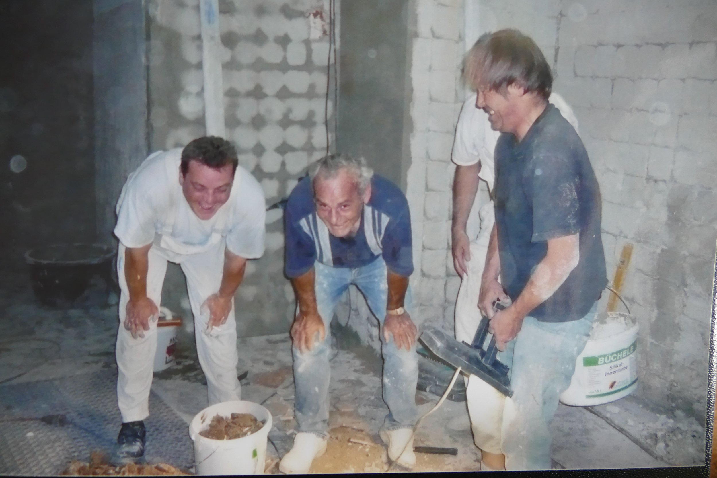 Vier Männer beim Bau eines Raumes, einer hält eine Blasmaschine, andere bücken sich bei Baumarbeiten, im Hintergrund Ziegelwände und Bauwerkzeuge.