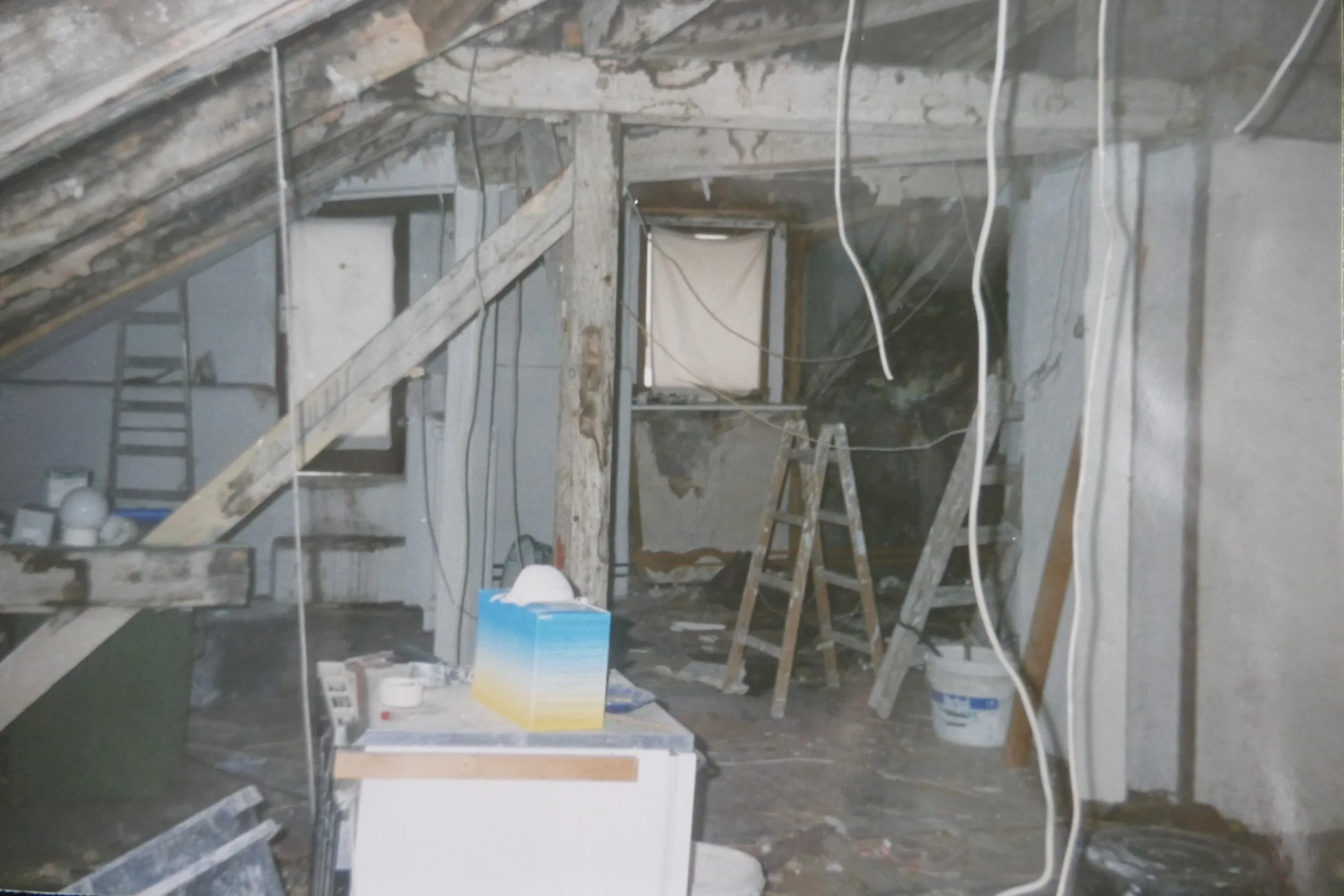 Ein Dachboden im Umbau mit sichtbaren Holzbalken, Kabeln, einer Leiter und verschiedenen Baumaterialien wie Putz und Werkzeugen.