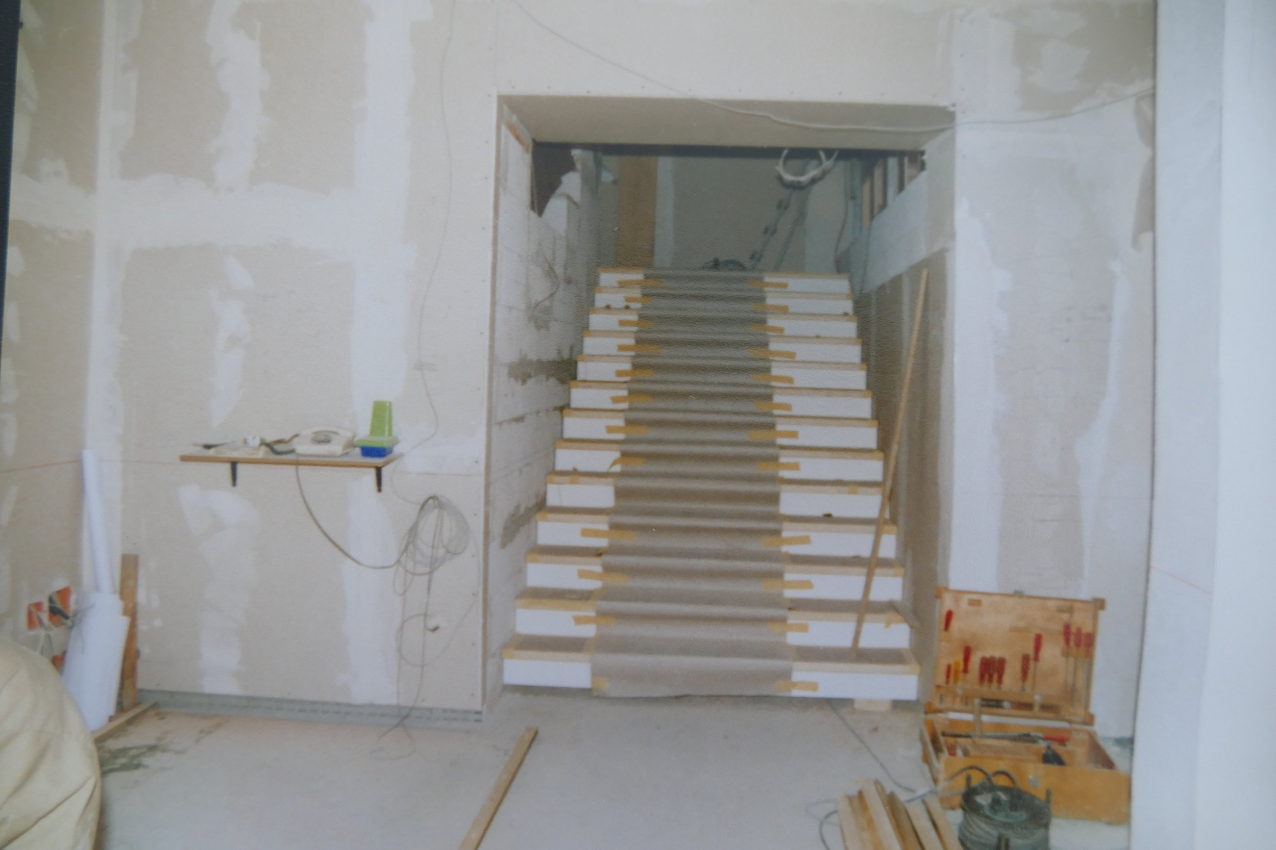 Innenansicht eines Raumes im Bau, mit einer im Bau befindlichen Treppe aus Holz, unvollständiger Wandverkleidung und Baumaterialien.