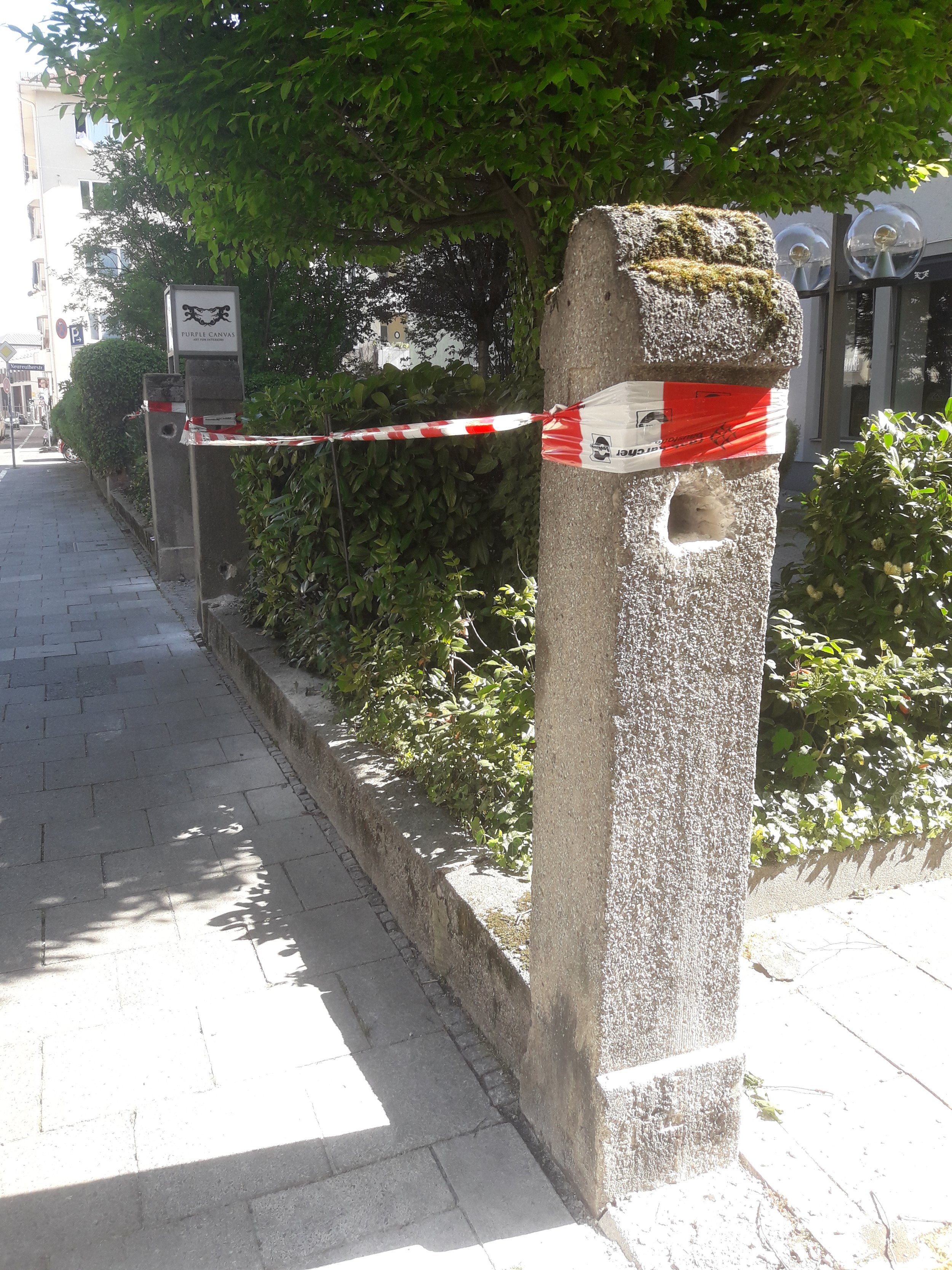 Ein Schild mit der Aufschrift "Purple Canvas" und einem schwarzen Logo steht neben einem großen, mit Moos bewachsenen Stein, der mit rotem und weißem Absperrband umwickelt ist, vor einem Garten mit Büschen und Bäumen auf einer Stadtstraße.