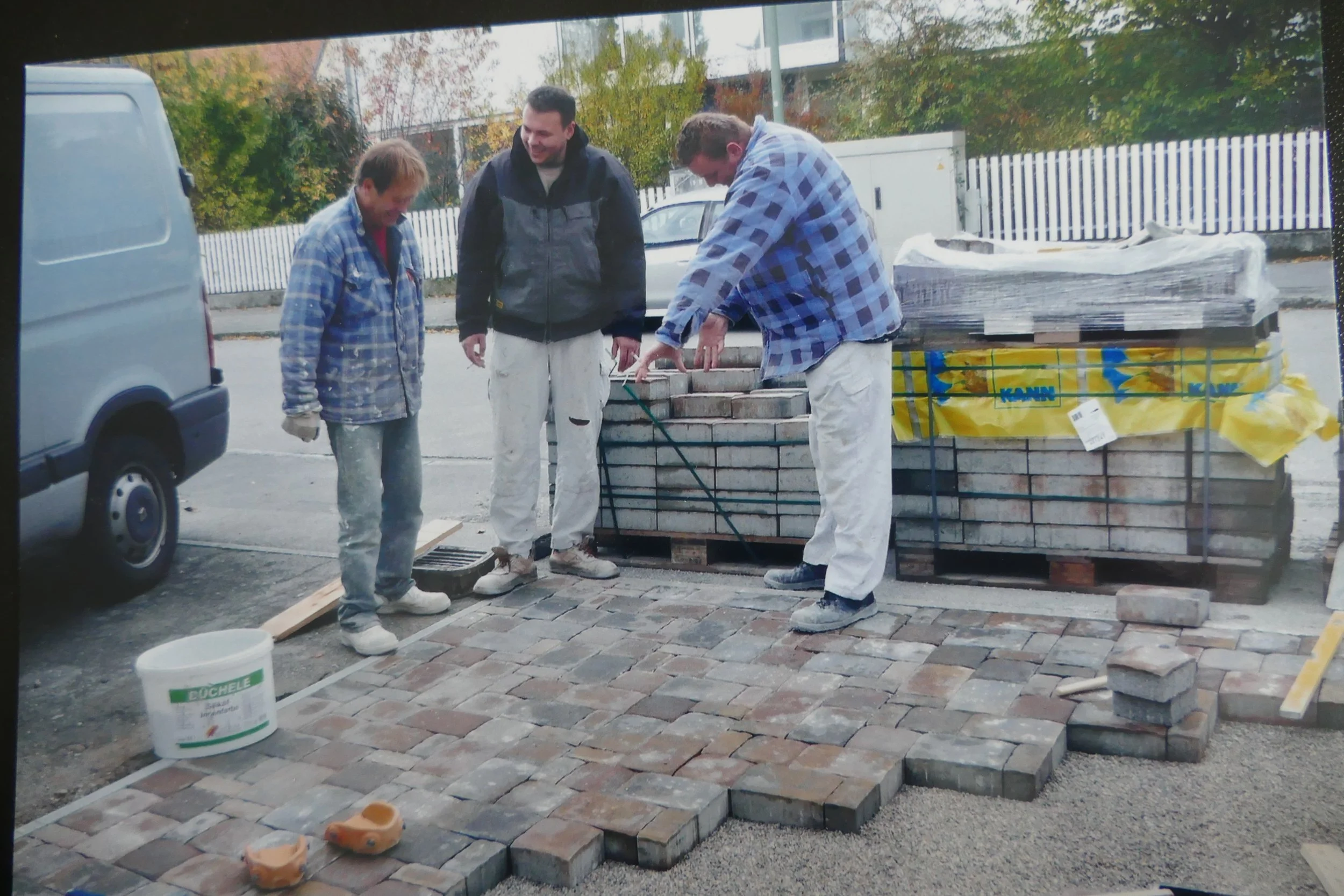 Drei Männer arbeiten an einer Pflastersteinverlegung im Außenbereich, während sie auf der vorhandenen Pflasterfläche stehen, um das Gebiet zu inspizieren. Es gibt einen Stapel Pflastersteine, eine Farbdose und andere Werkzeuge auf der Baustelle.