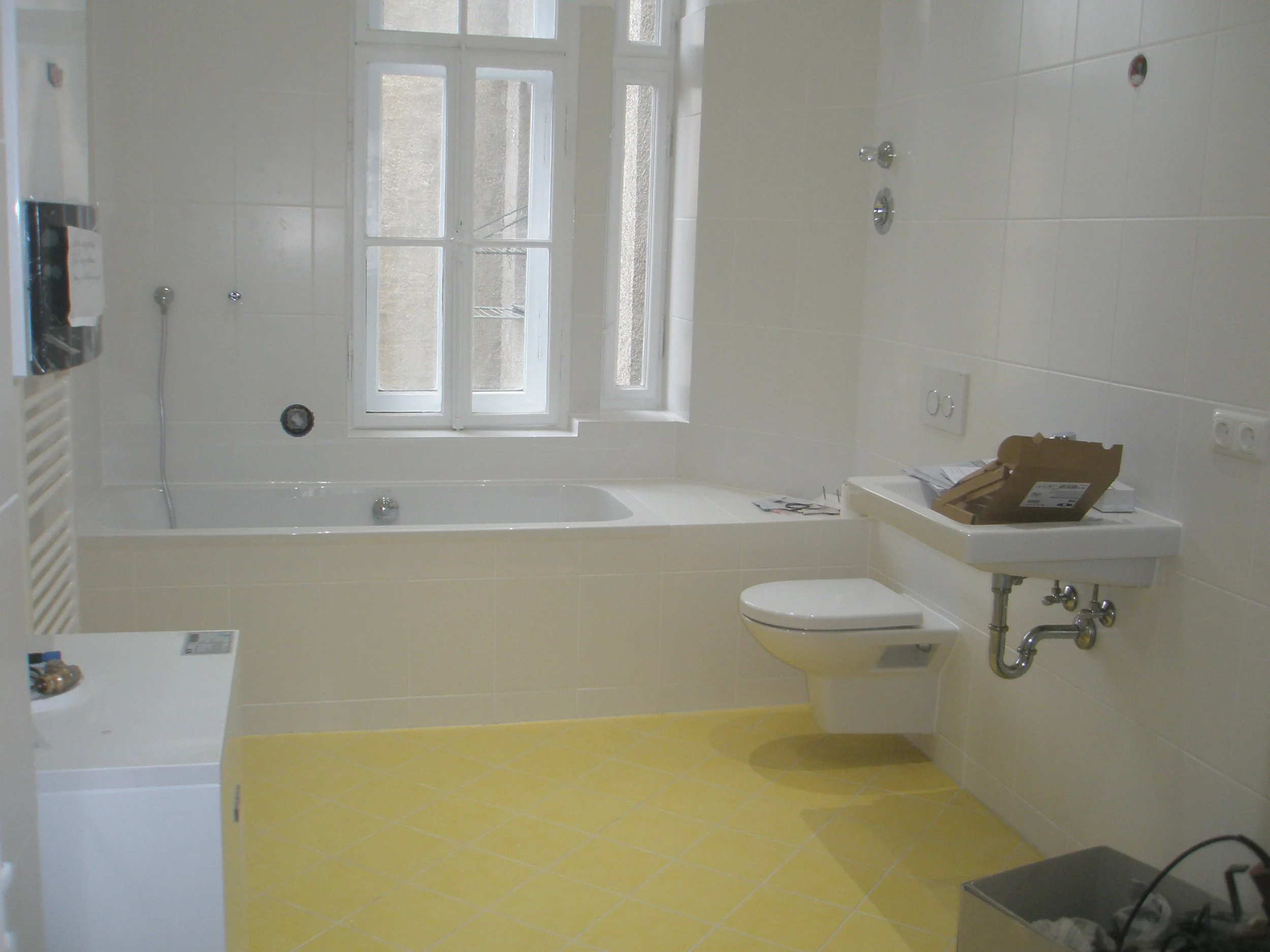 Innenansicht eines unfertigen Badezimmers mit weißem Fliesen, einer Badewanne, einem Waschbecken, einer großen Fensterscheibe, und noch installiertem Wasserhahn sowie elektrische Steckdosen und einem geöffneten Karton auf dem Waschtisch.