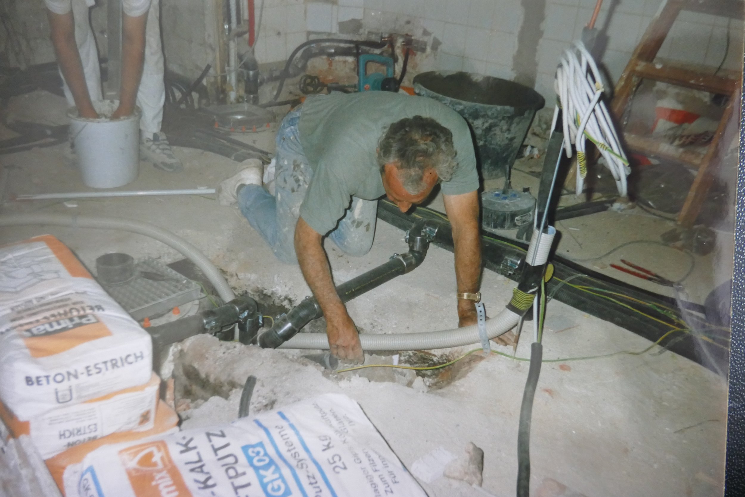 Mann arbeitet in einem unordentlichen Keller, umgeben von Bau- und Sanitärmaterialien, Kabeln und Werkzeugen.