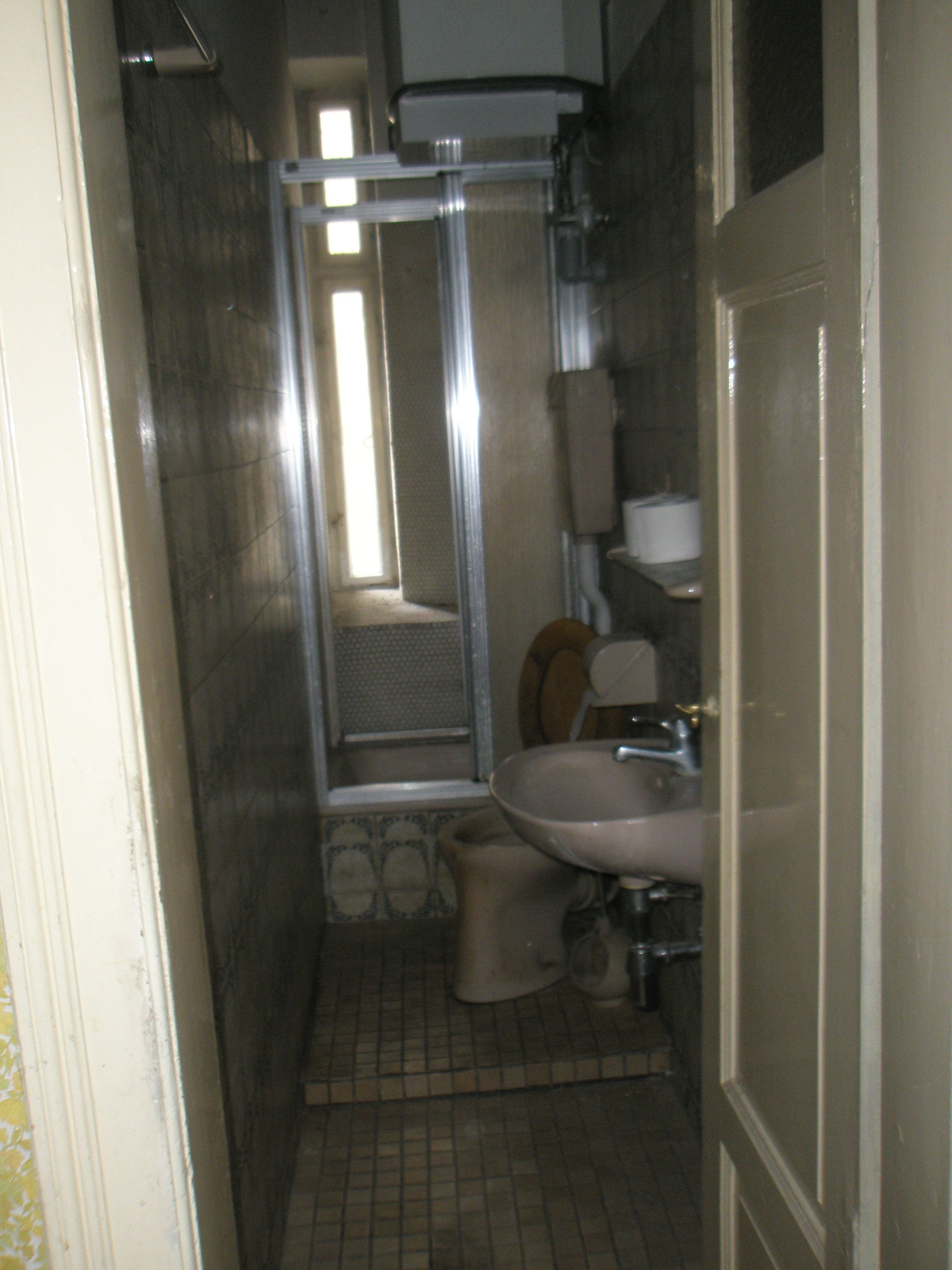 Ein veraltetes, kleines Badezimmer mit schwarzem Boden, einer Toilette, einem Waschbecken und einer Duschkabine.