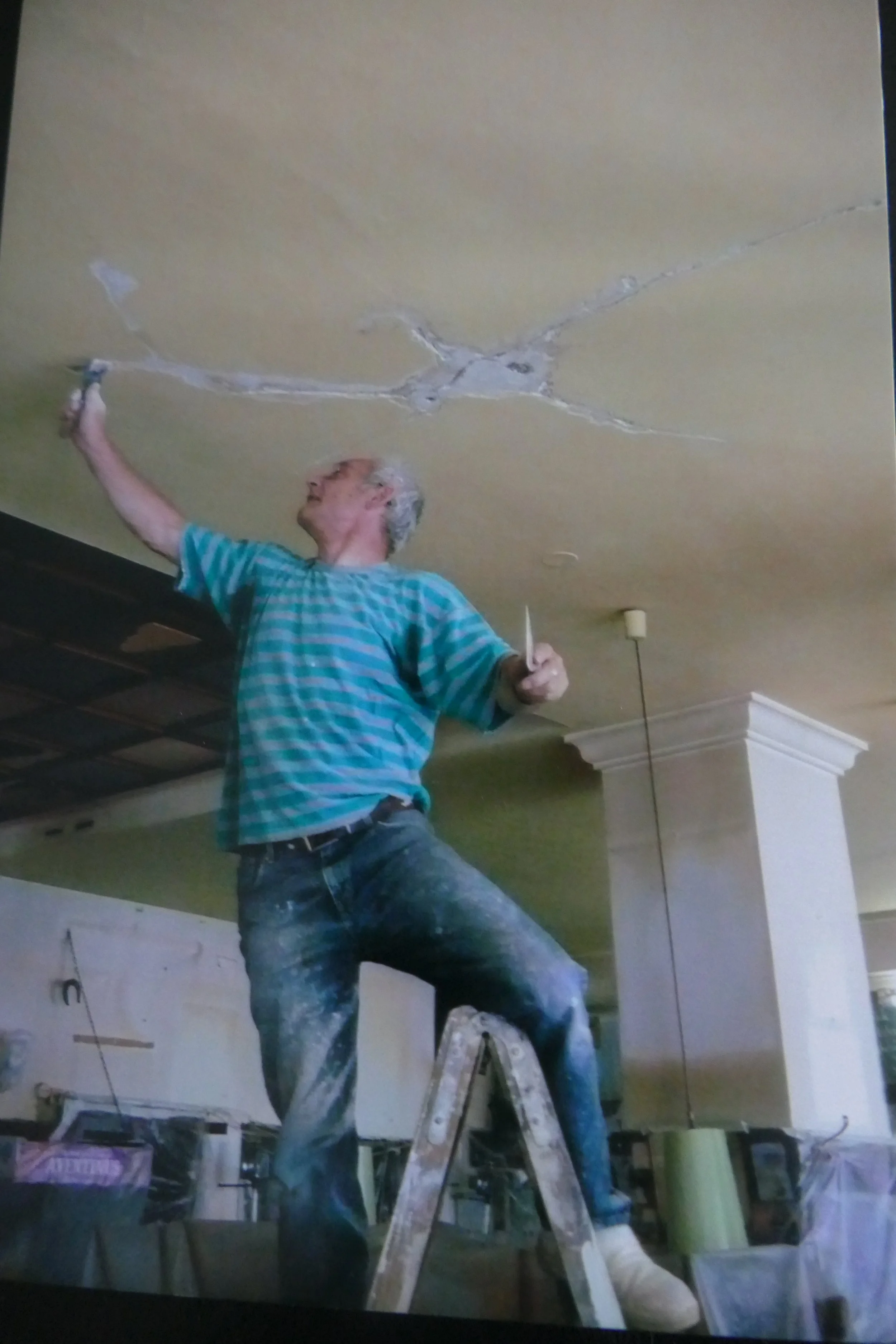 Ein Mann steht auf einer Leiter und repariert die Decke eines Zimmers. Er trägt eine blau-karierte Hemd und Jeans, scheint zu streichen oder zu reparieren.