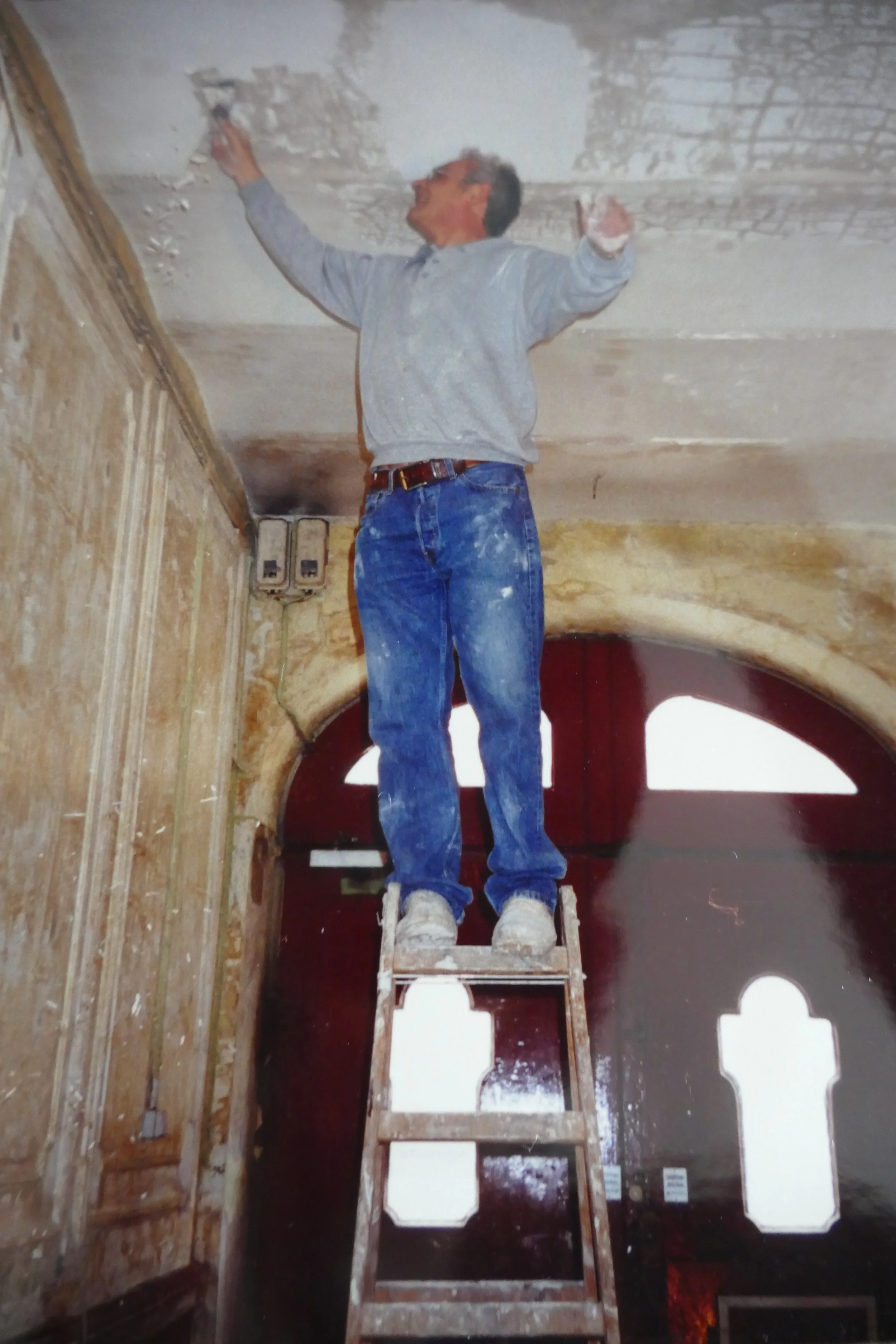 Ein Mann steht auf einer Leiter in einem Raum mit abgeblätterter Decke und alten Wänden und arbeitet an der Decke oder Wand. Er trägt eine graue Sweatshirt und blaue Jeans.