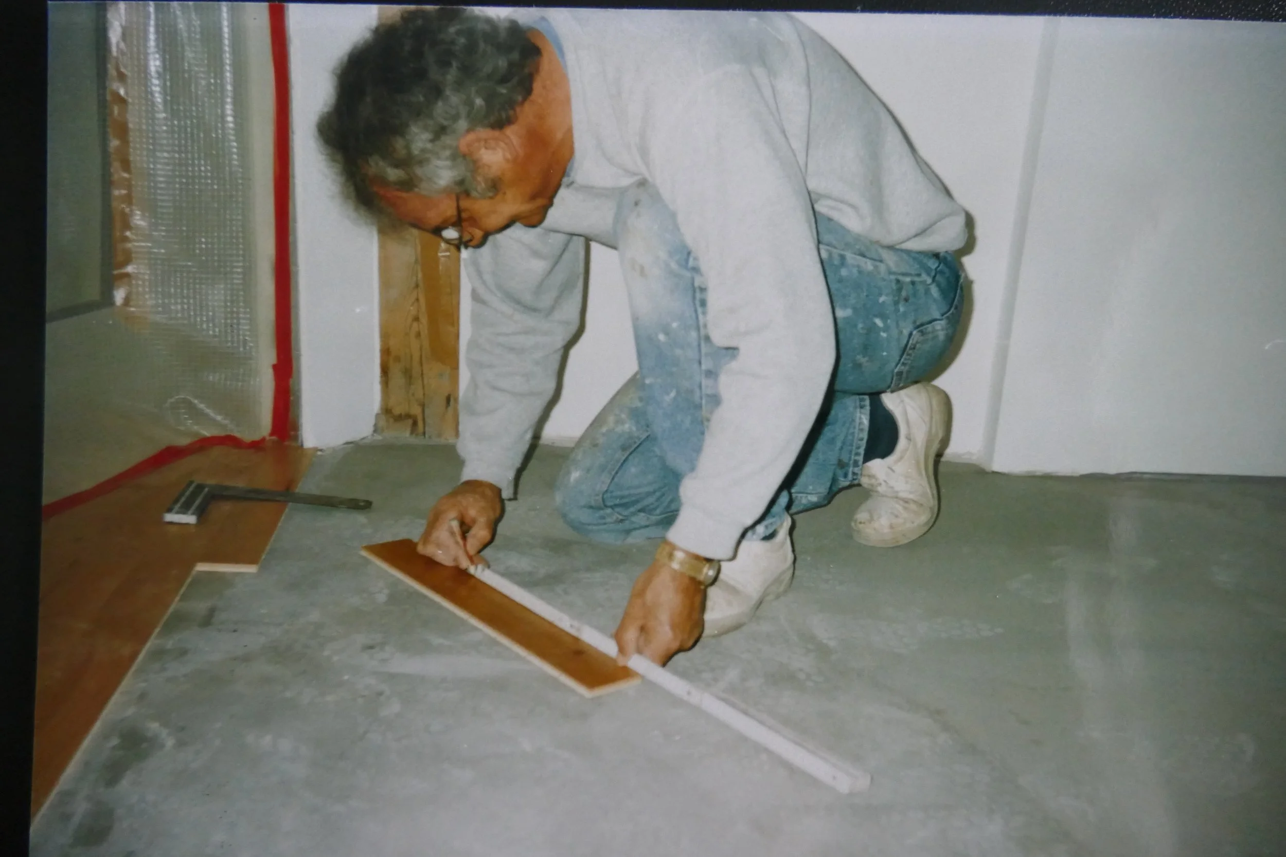 Älterer Mann kniet auf dem Boden, zeichnet mit einem Lineal eine Linie auf eine große Holzplatte, trägt weiße Schuhe, eine graue Jacke und Jeans, in einem Raum mit weißen Wänden.
