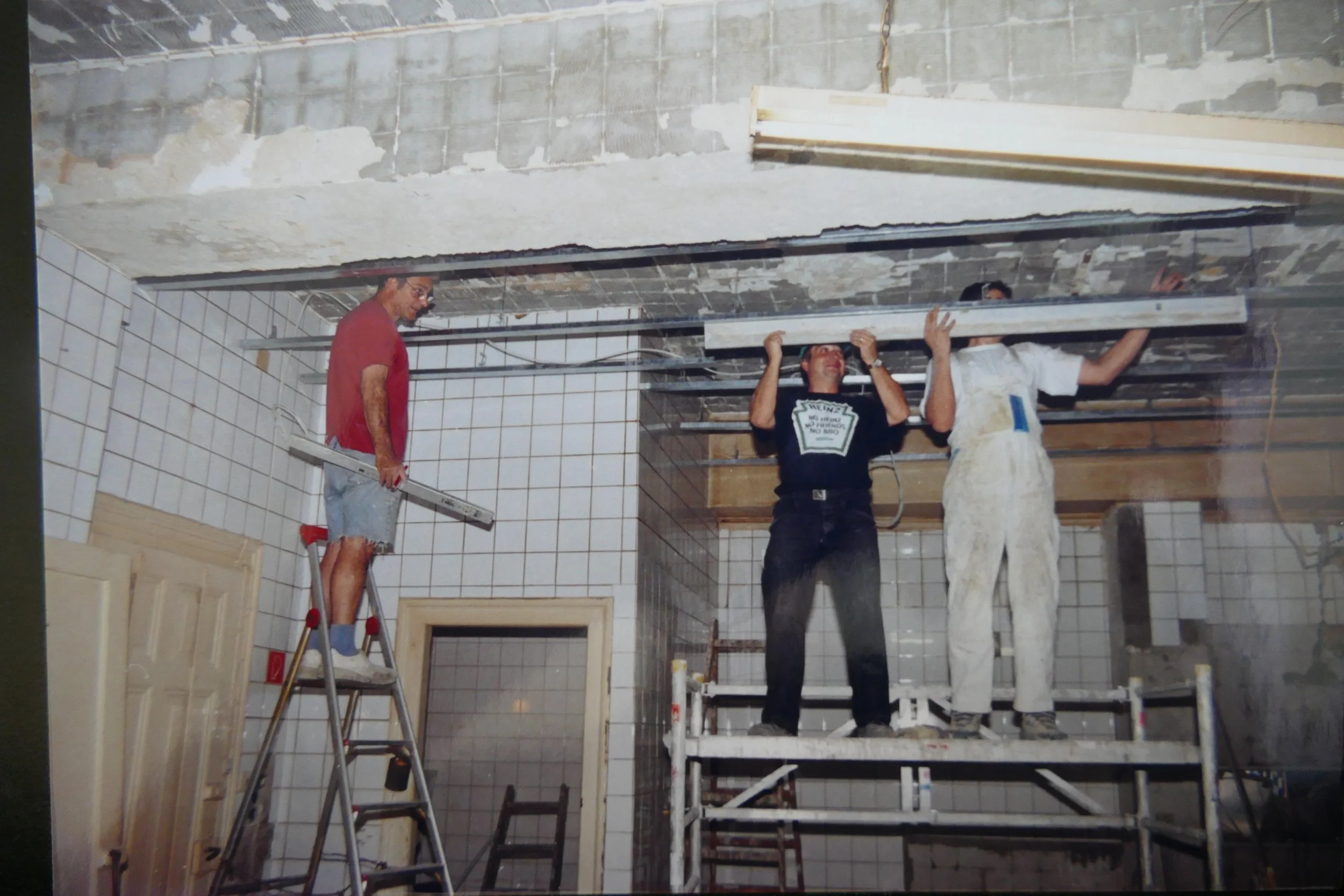 Drei Männer auf einer Baustelle in einem Raum mit Fliesen, die an der Decke und an den Wänden installiert werden. Zwei Männer stehen auf einer Gerüstbühne, einer steht auf einem Leiter. Sie arbeiten an der Decke.