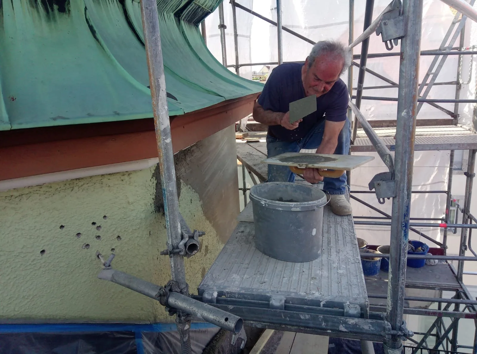 Mann bei Renovierungsarbeiten auf Gerüst, in der Nähe eines Gebäudes mit grün gestrichener Wand, wendet Putz oder Farbe auf die Fassade an.