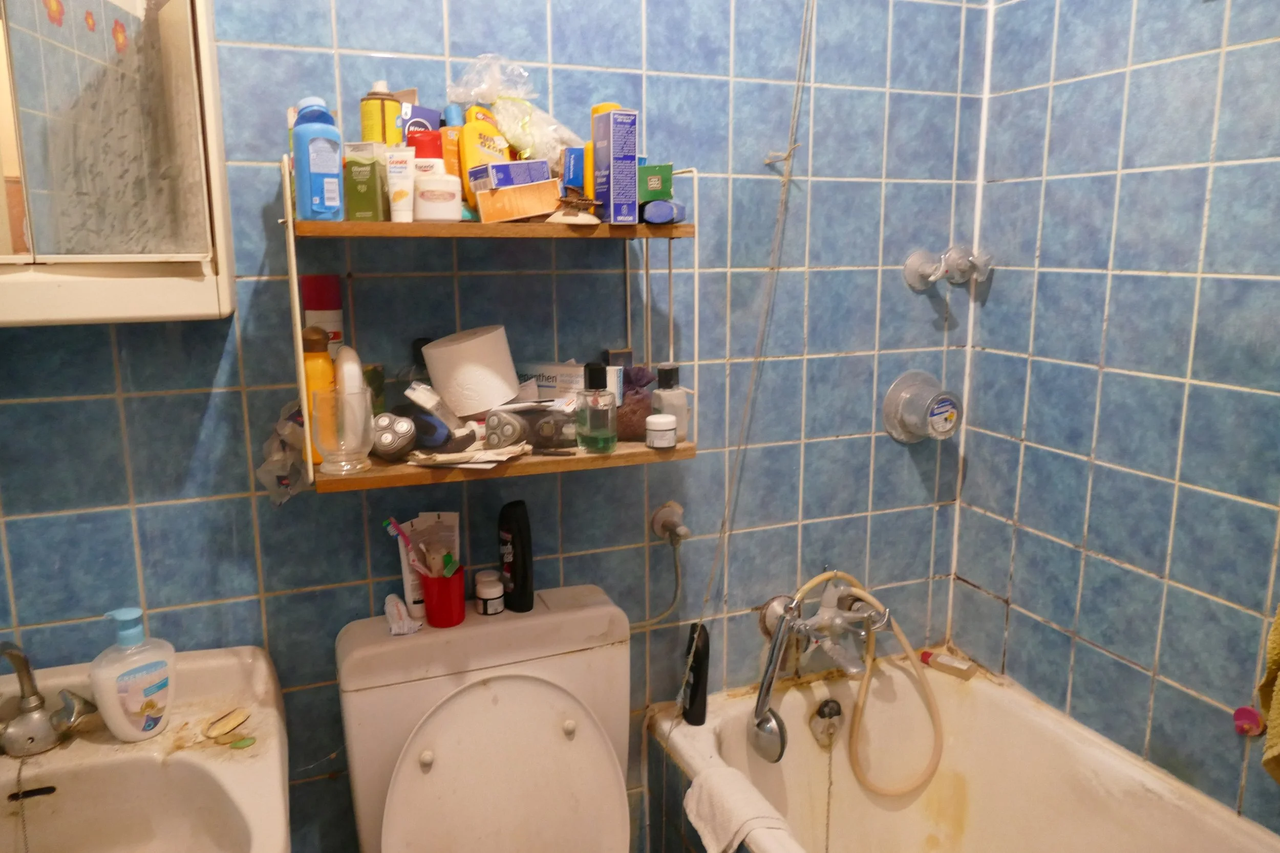 Ein schmutziges Badezimmer mit blauen Fliesen, in dem Badezimmerartikel, ein Staubsauger, eine Toilette, ein Waschbecken und ein Badewannenbereich mit Duschkopf, Wasserhähnen und ungeordneten Gegenständen sichtbar sind.