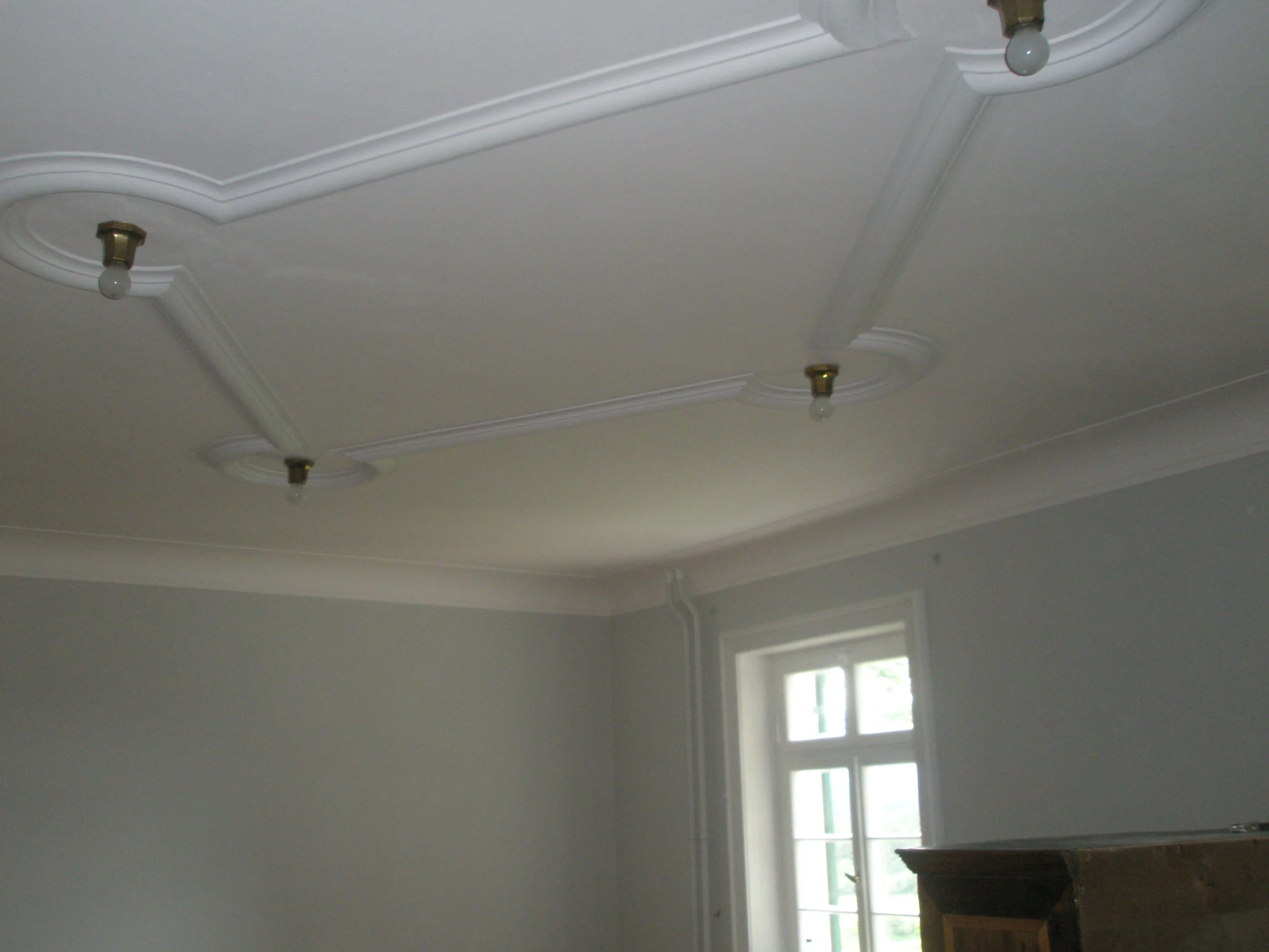 Decke mit weißen Verzierungen, Lampenfassung ohne Glühbirne, Fenster mit mehreren Scheiben, viel natürliches Licht in einem Raum.